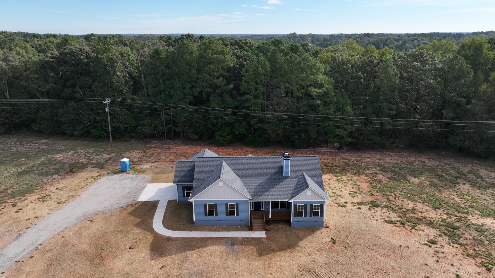 Blue-roofed house with black shuttered windows, wooden stairs leading to entry, driveway, grassy yard, and mature trees with visible power lines in the background