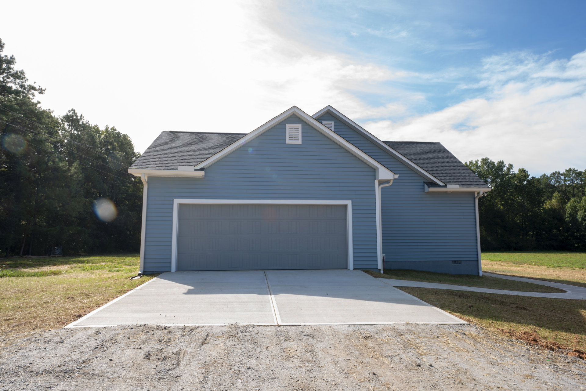 Blue-sided custom home with white-trimmed garage door, concrete driveway, white vent, overhead power lines, and mature trees under partly cloudy sky