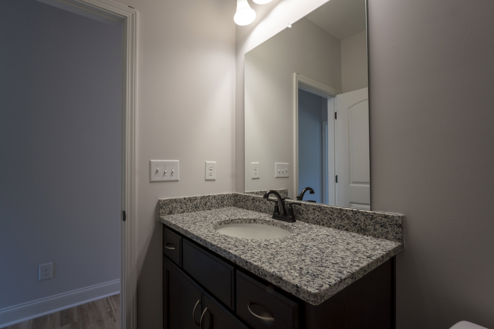 Marble countertop vanity with undermount sink, chrome faucet, large framed mirror, white tile backsplash, electrical outlet, row of wall switches, white door with black handle