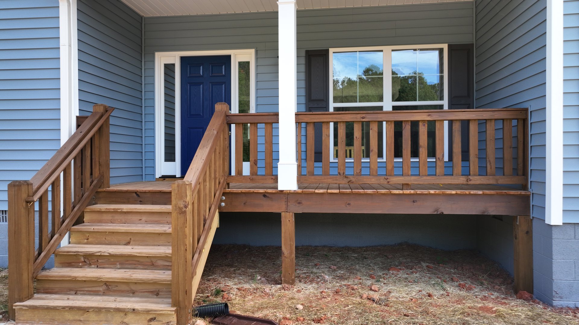 Wooden deck with white columns and railings, blue front door with white trim, large window reflecting trees, wood posts supporting covered porch, ground covered in hay and rocks