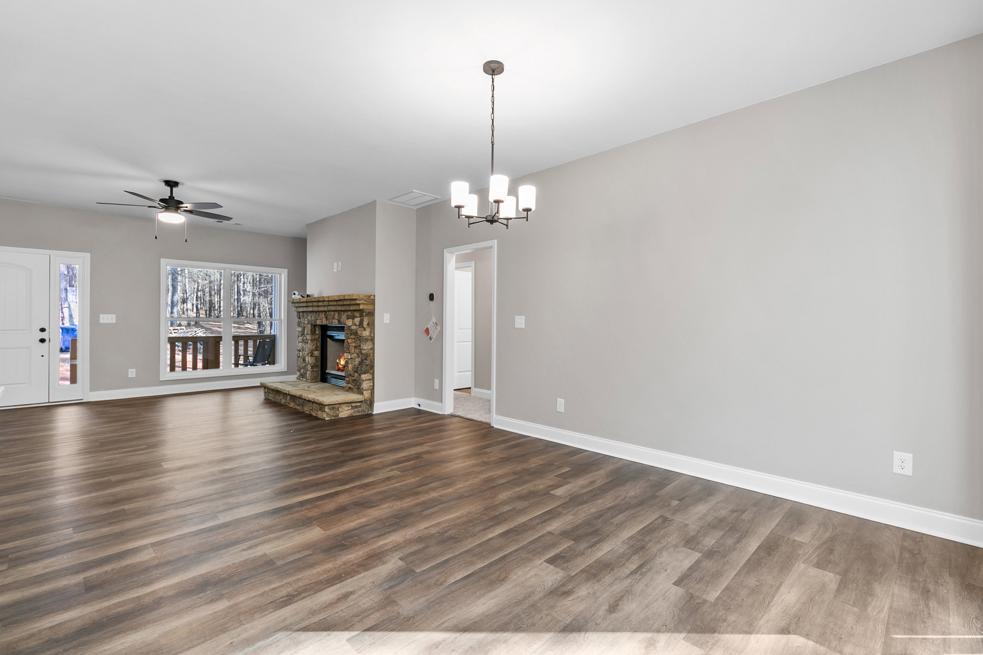 Living room with hardwood flooring, stone fireplace with burning fire, large window overlooking deck with outdoor chair, modern ceiling fan and light fixture, white plaster walls