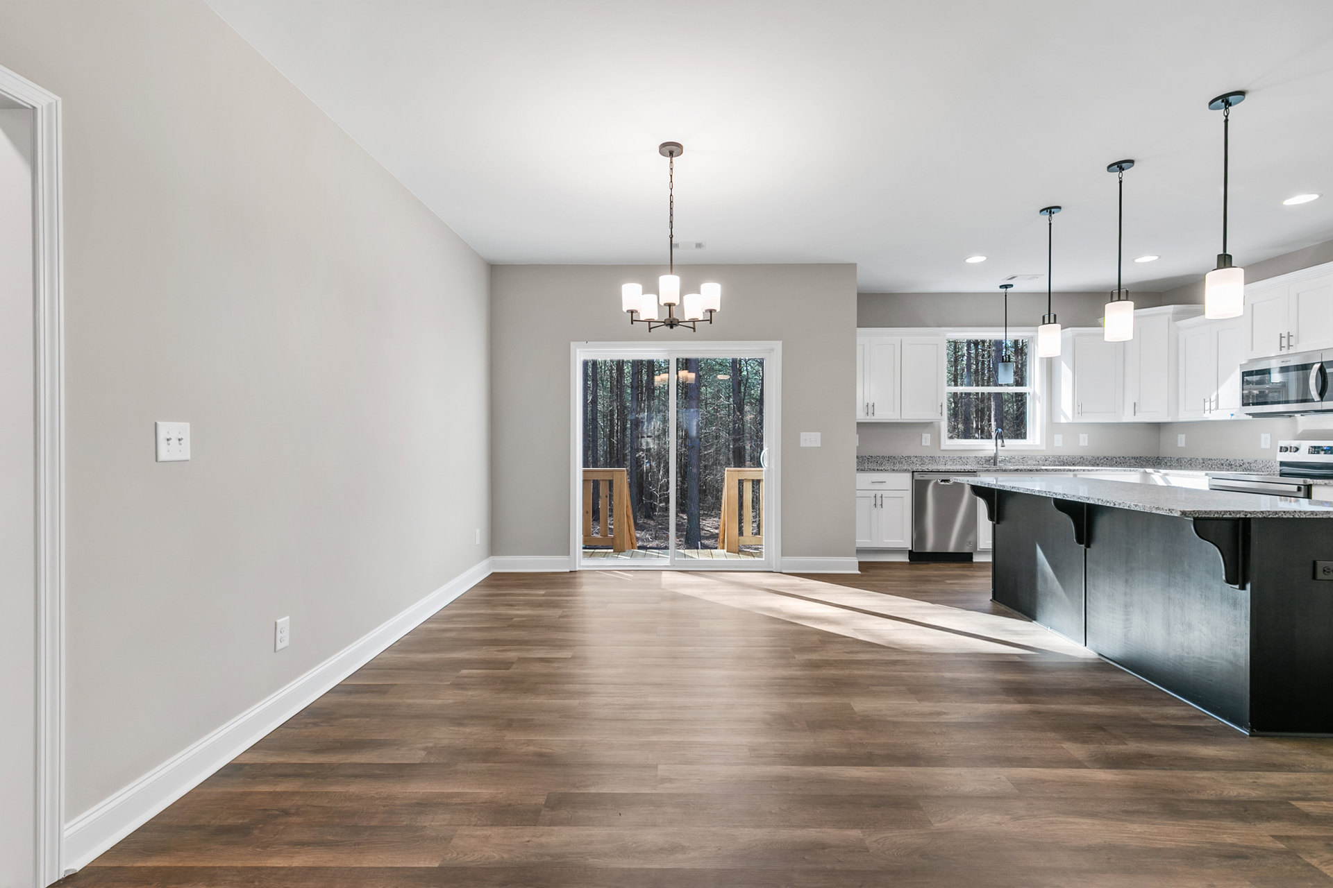 Open kitchen and dining area featuring wood flooring, black marble countertop, white cabinetry, sliding glass door overlooking wooded landscape, modern light fixture with white