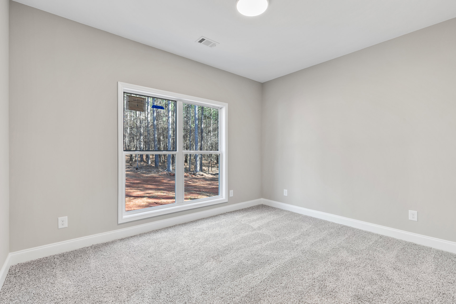 Neutral-toned carpeted room with white walls, large window overlooking trees, ceiling vent and recessed light, newspaper resting on window sill, subtle baseboard molding