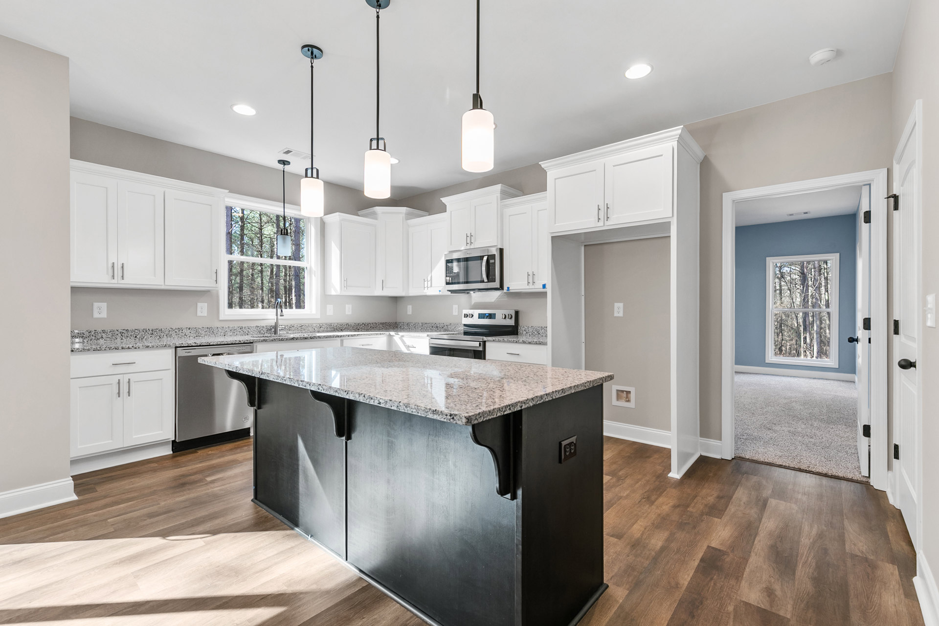 Kitchen featuring a central island with granite countertop, white shaker cabinets, stainless steel microwave, undermount sink, pendant lighting, and window overlooking trees.