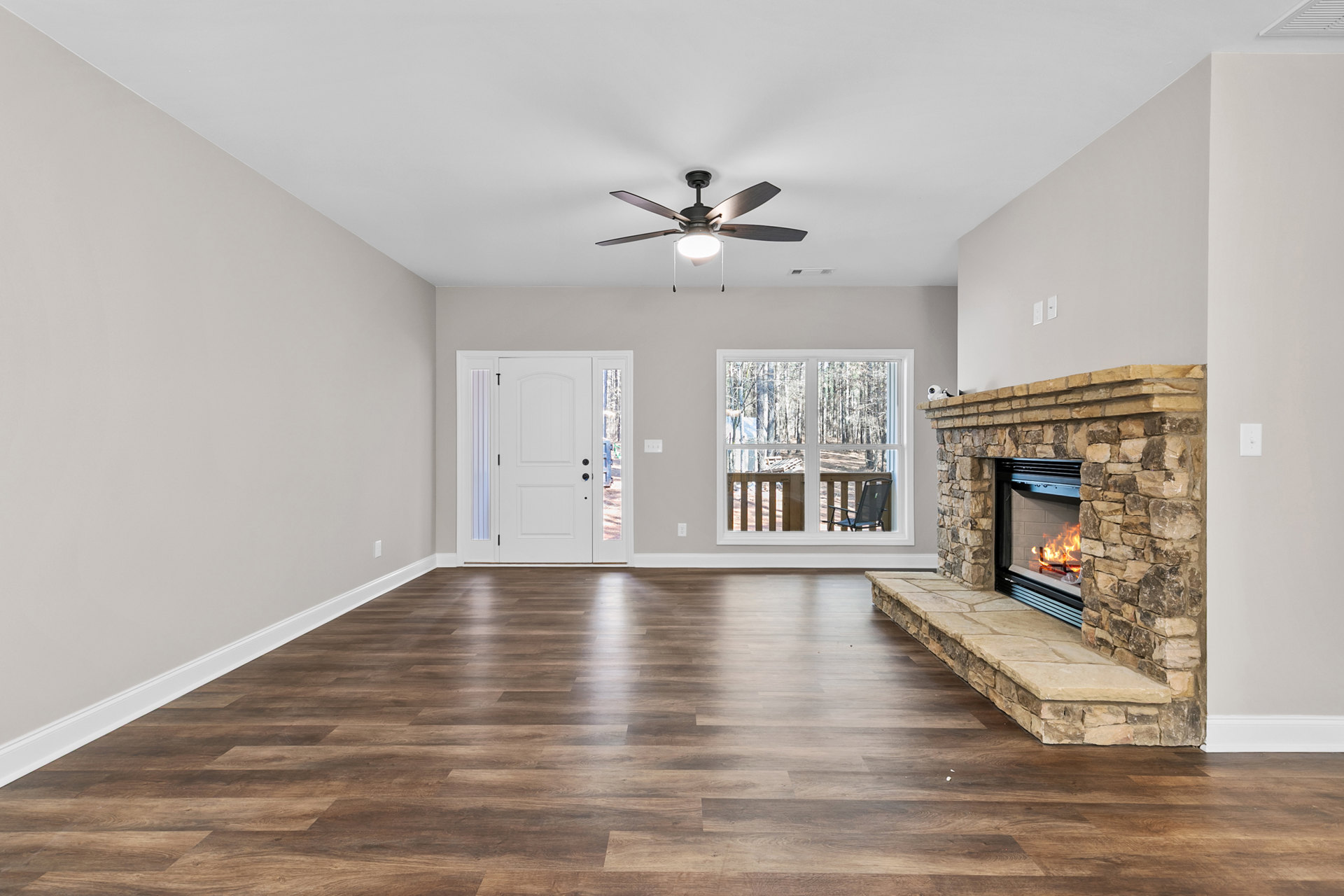 Living room with laminate wood flooring, plaster walls, central fireplace with white mantel, ceiling fan with light, glass-panel door, and window showing outdoor chair.