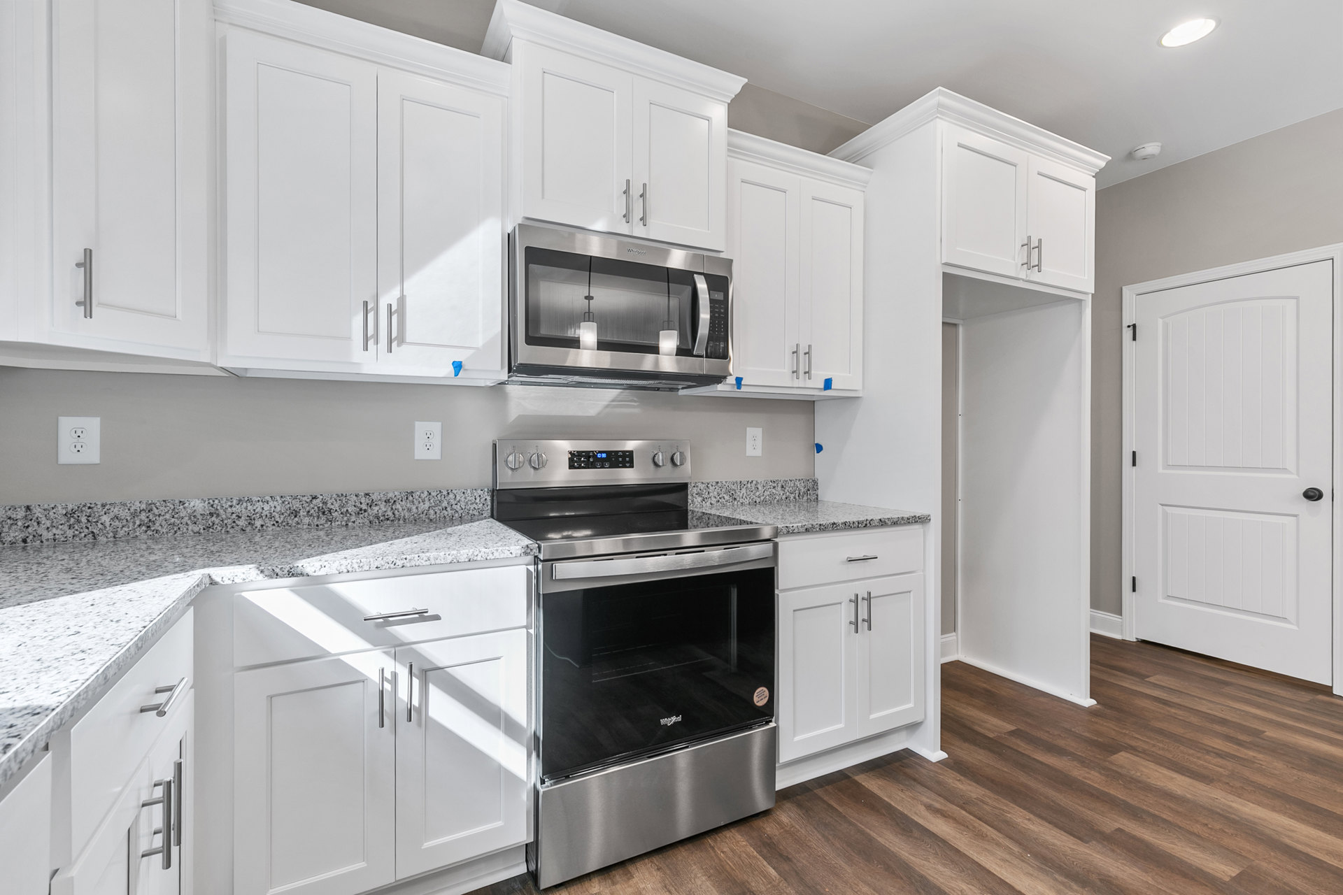 White kitchen cabinets, stainless steel oven with black glass door, built-in microwave above countertop, white door with black handle, electrical outlet, and modern ceiling light