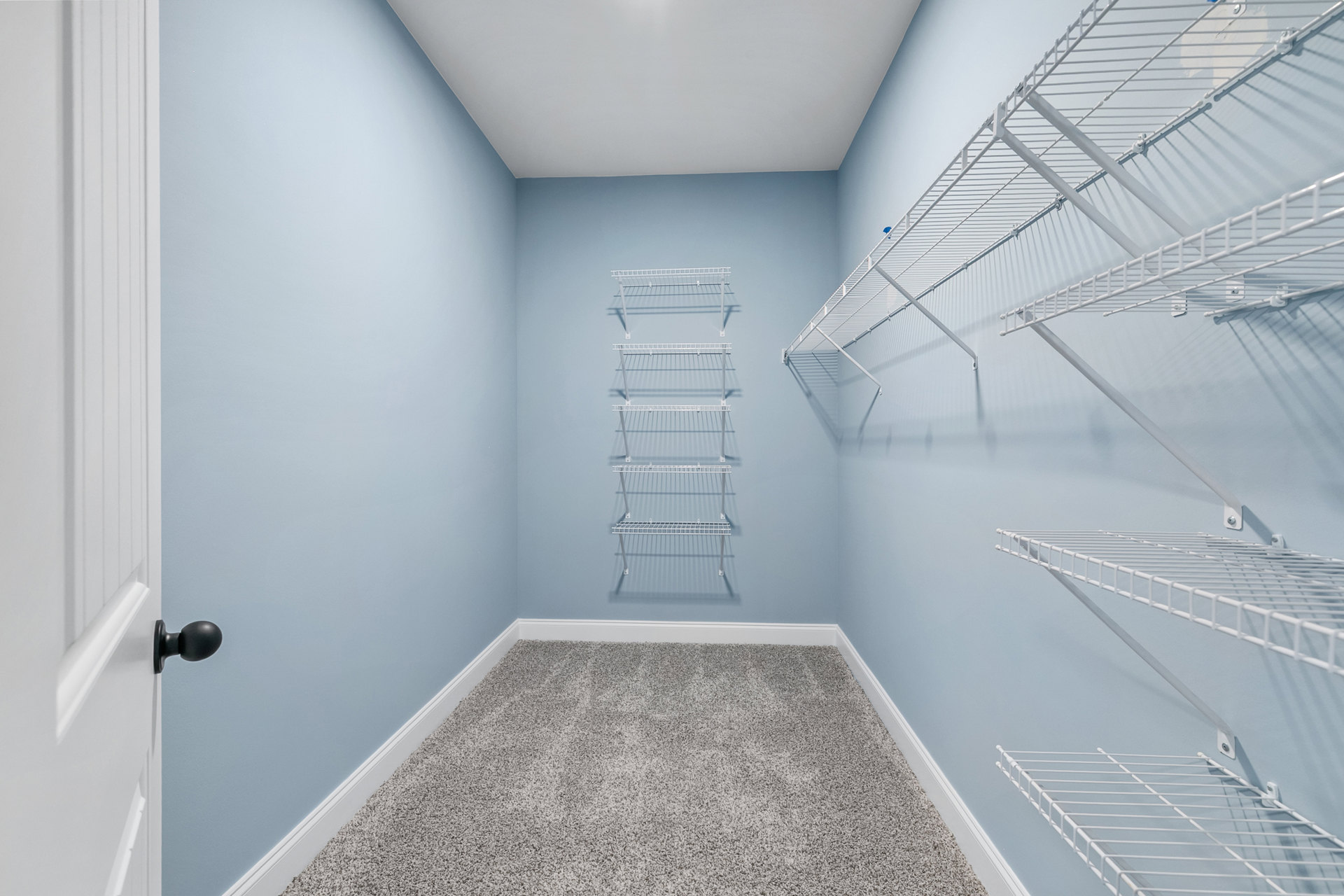 White built-in shelves mounted on a plaster wall above beige carpet flooring, with a white clothes rack and a close-up of a brushed metal door knob visible.