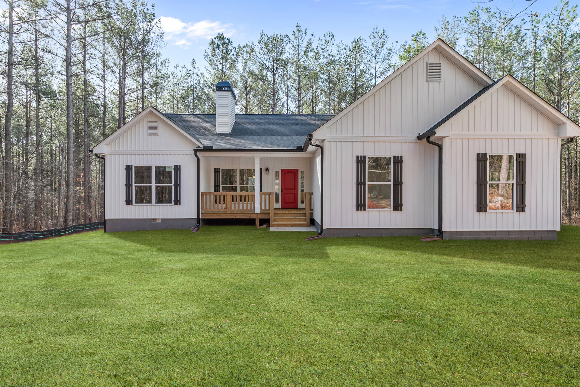 White siding house with red front door, white trim, wooden porch railing, green lawn, and large windows; trees and clouds in background