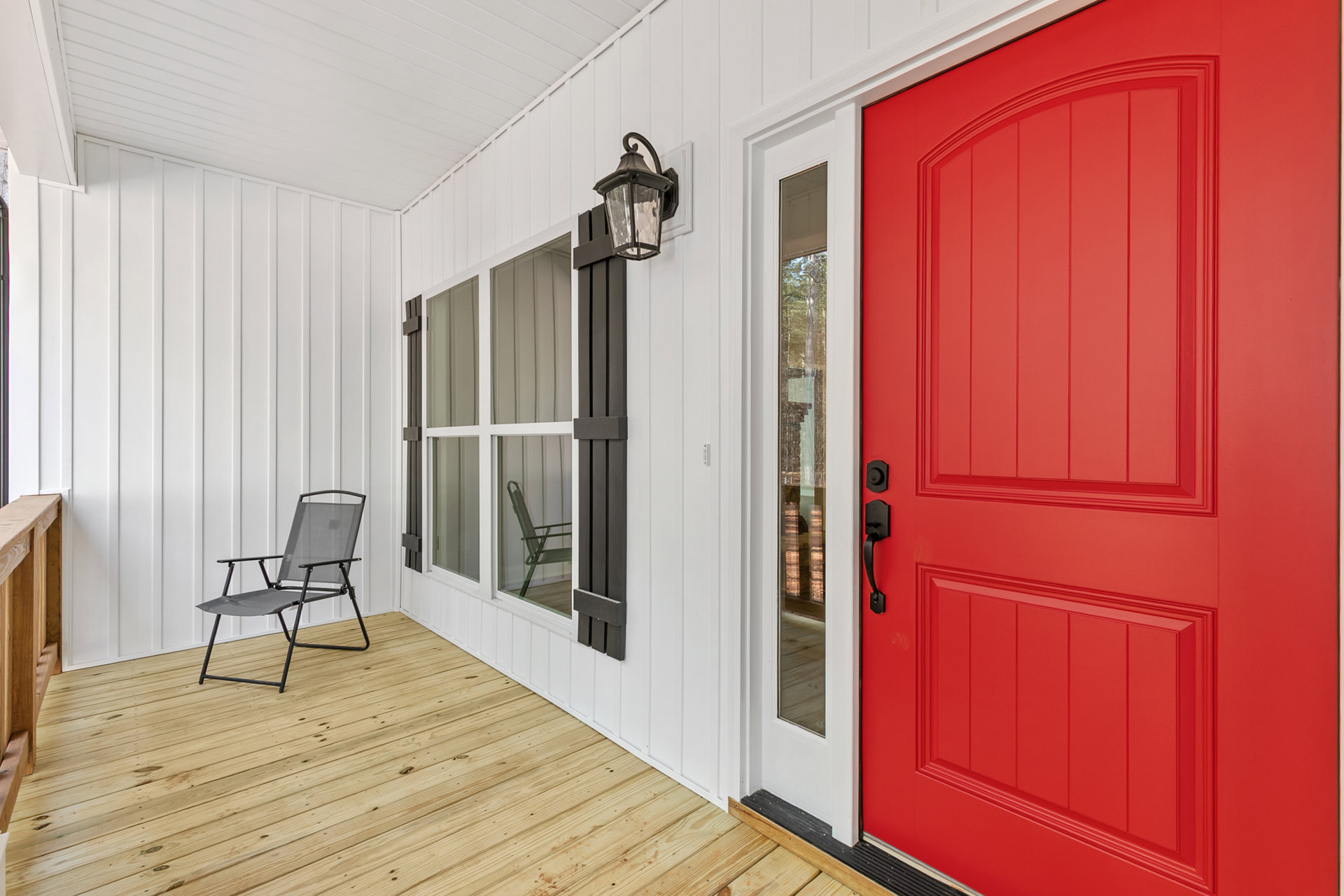 Red front door with black handle, wooden porch floor, single chair beside window, wall-mounted lamp above seating area.
