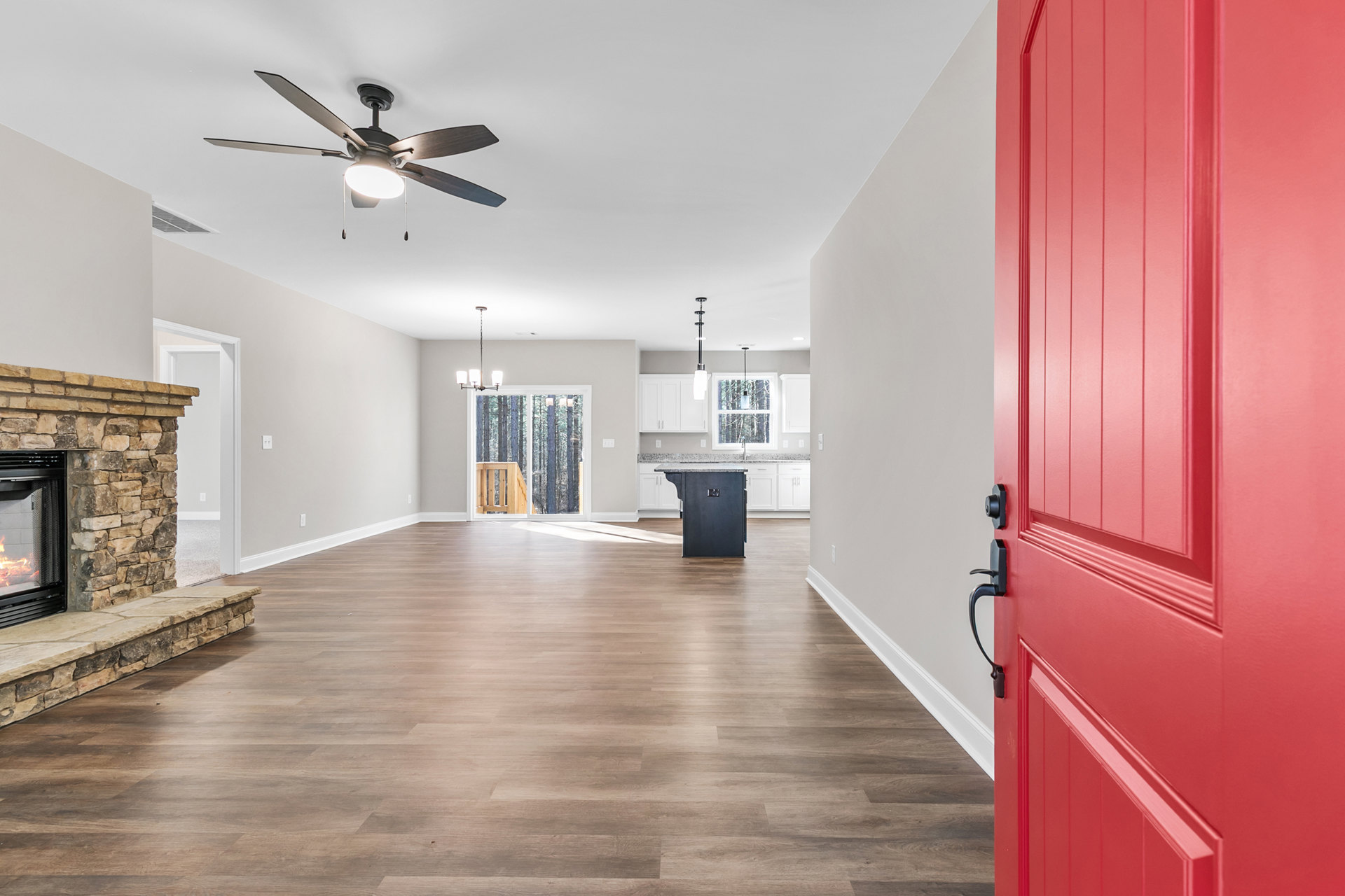 Red front door with brushed metal hardware, wood flooring, ceiling fan with light, stone fireplace with active fire, sliding glass door opening to wood deck.