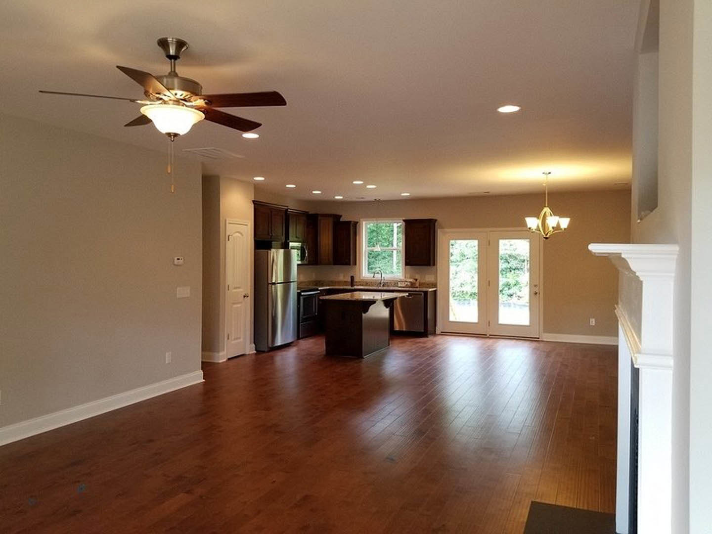 Open-concept kitchen and living room featuring wood laminate flooring, ceiling fan with light fixture, stainless steel refrigerator, glass-paneled double doors, dining table, and