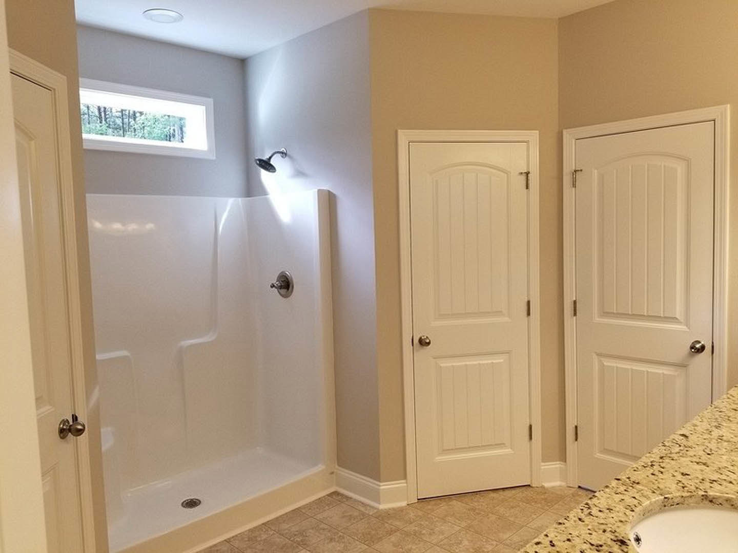 Modern bathroom featuring a glass-enclosed shower, white vanity with chrome faucet, white paneled door with silver handle, and window overlooking leafy trees