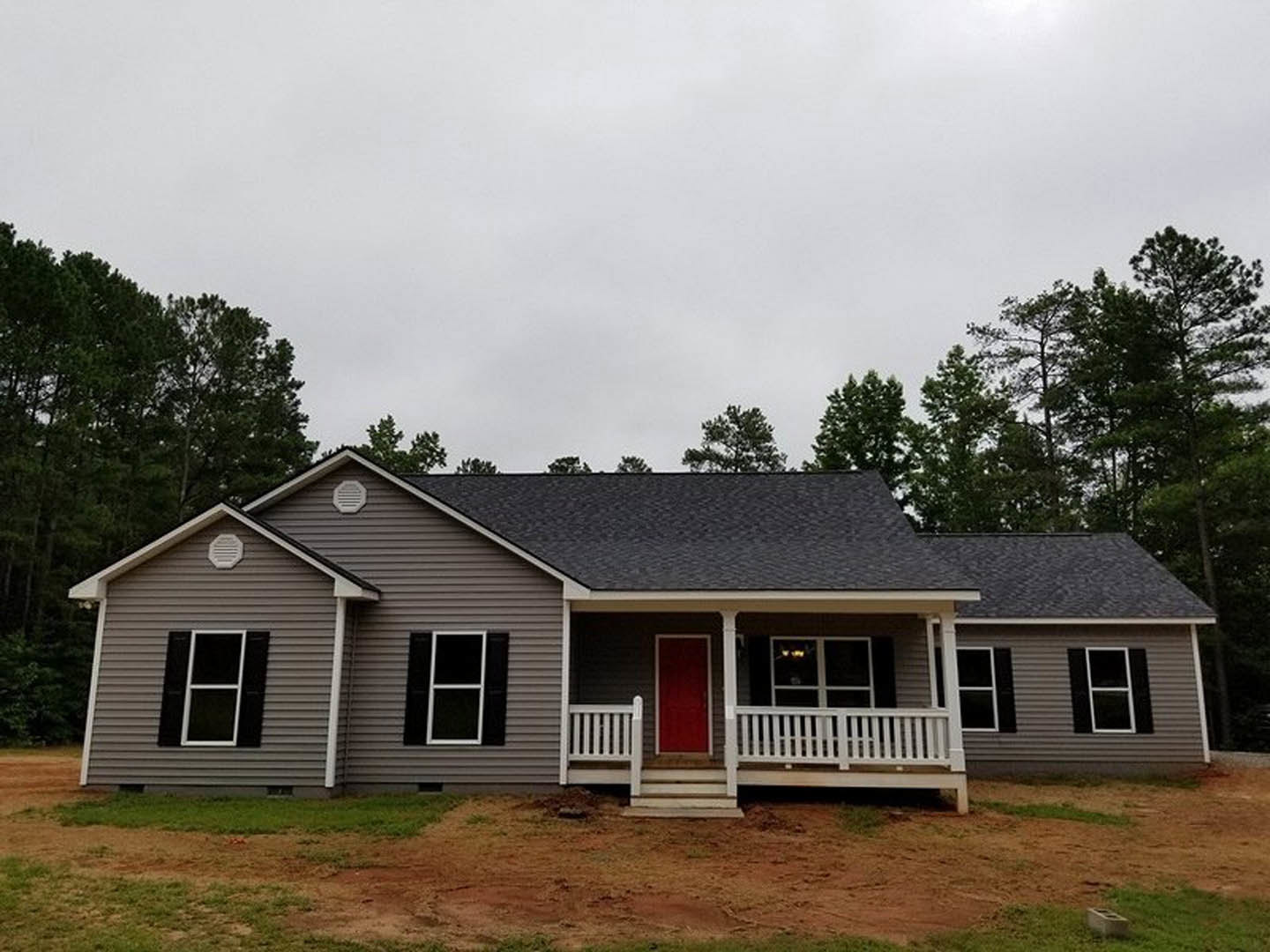Red front door with white frame, white porch railing, black shuttered window, white-framed window, dirt patch in front yard, cloudy sky above residential home.