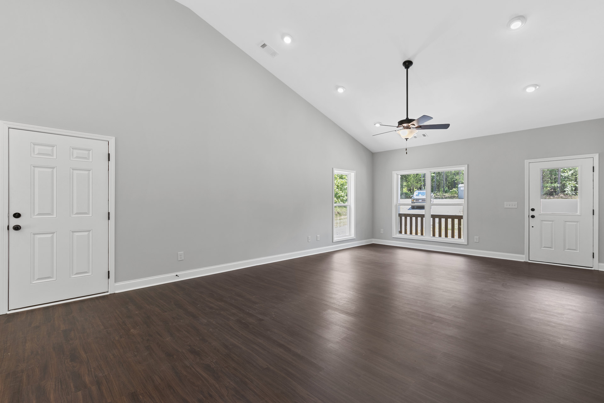 Spacious room with hardwood flooring, ceiling fan, white doors featuring black knobs and window panes, large window overlooking trees, partial view of driveway with parked car