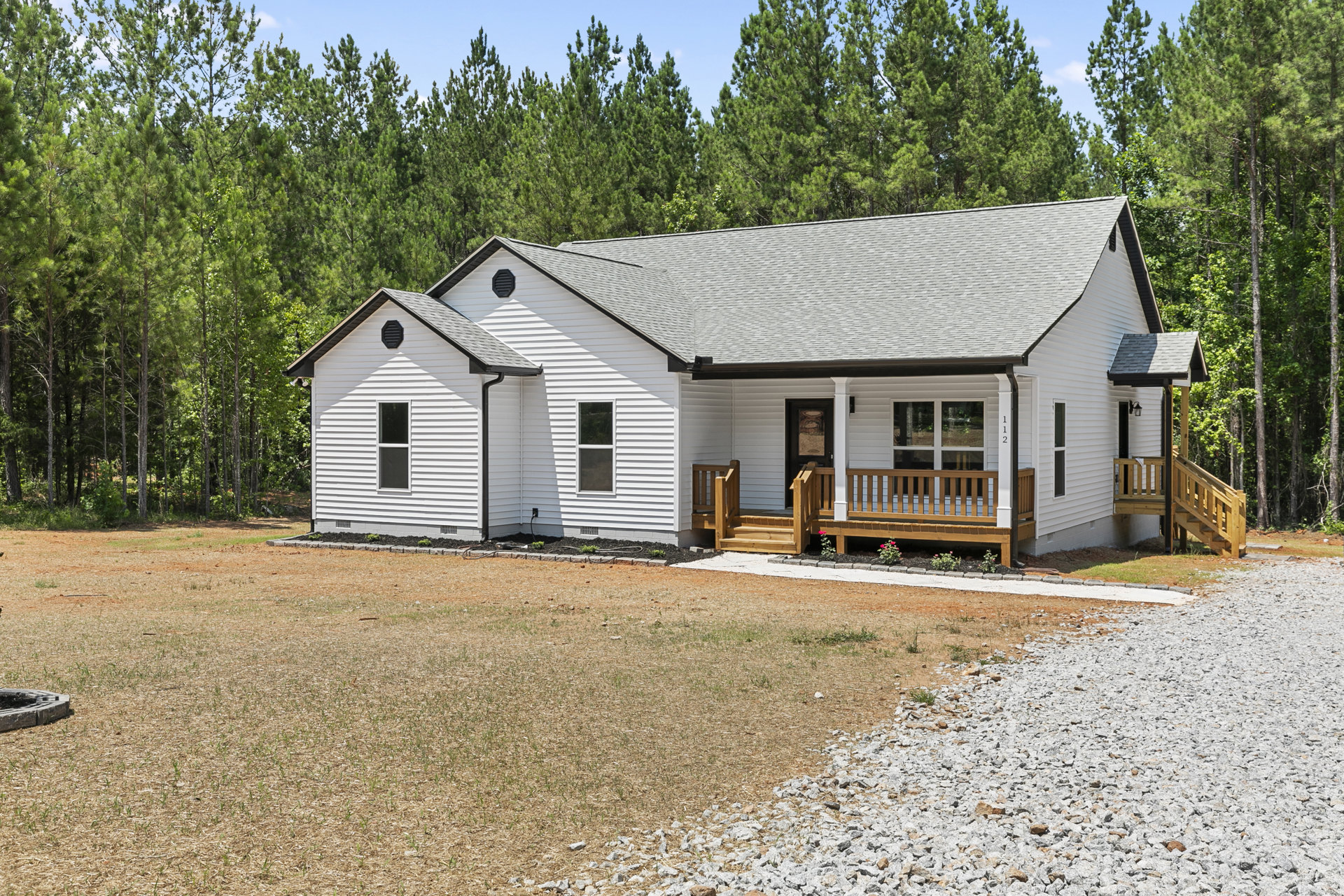 White house with black roof, gravel driveway, wood porch and deck, wooden bench with flowers, surrounded by trees and yard