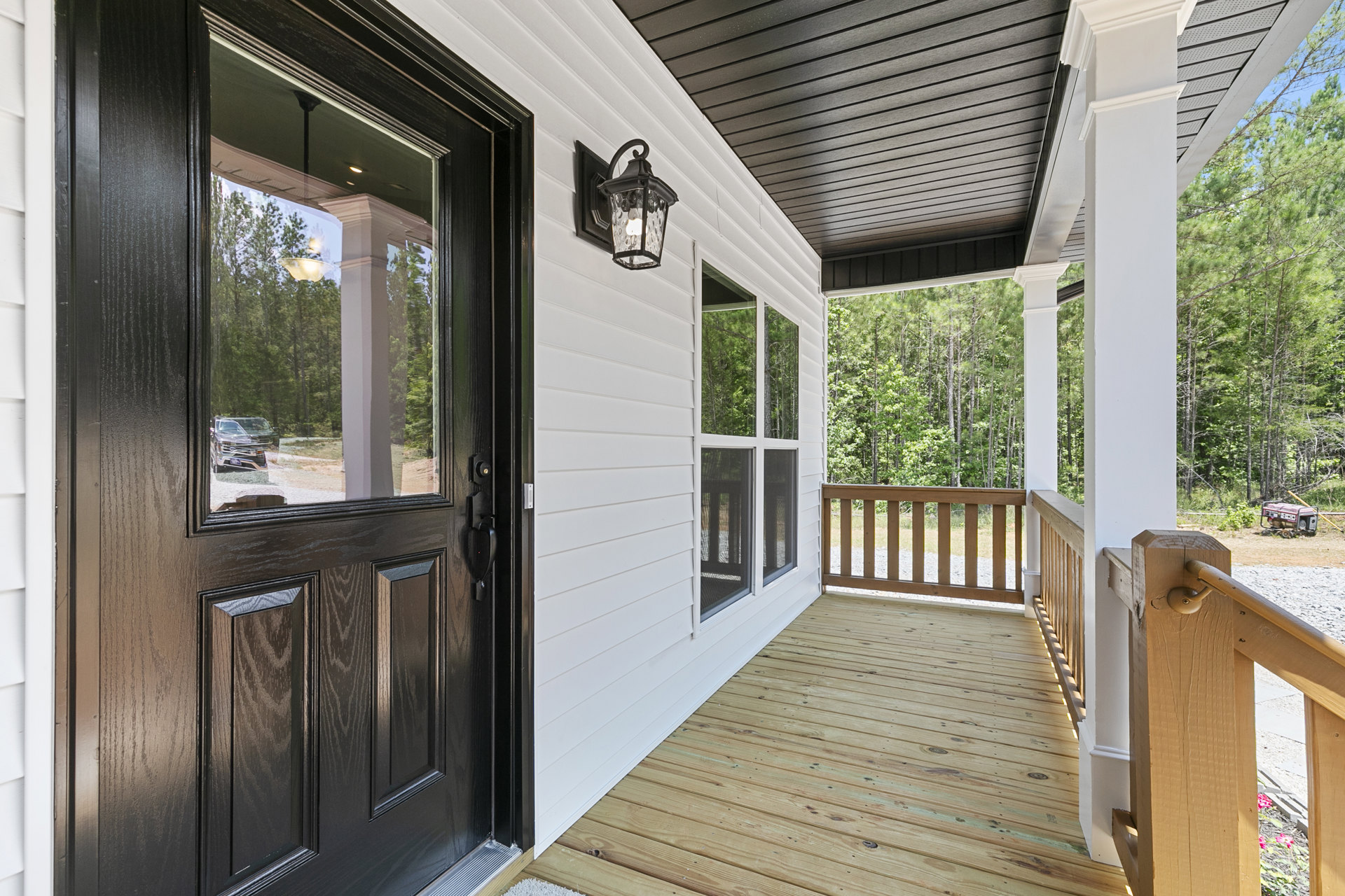 Wooden front porch with horizontal railing, glass door reflecting parked car, light fixture above entry, wide plank deck, and windows framed by painted siding.