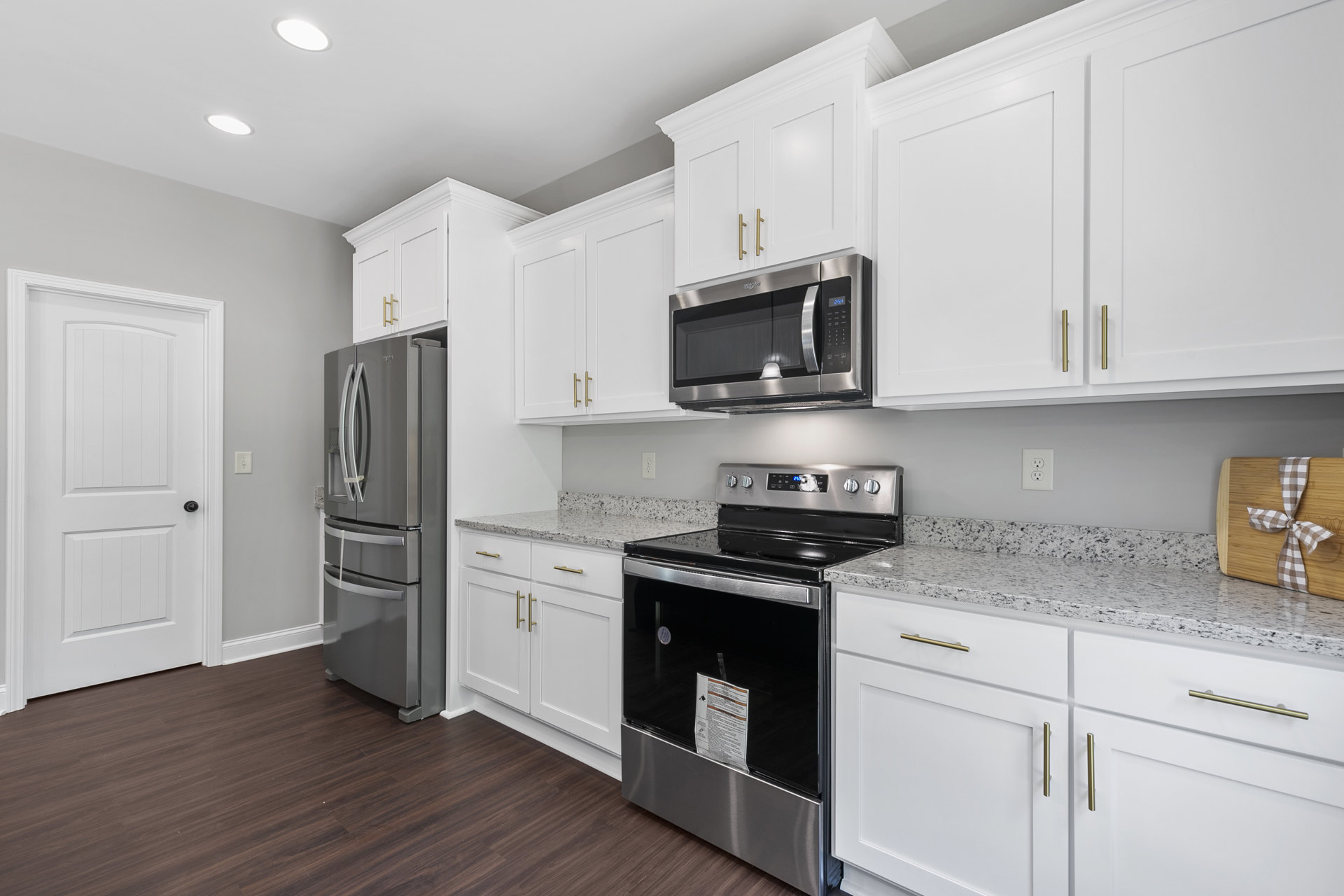 White kitchen with shaker cabinets, stainless steel refrigerator, stove with black door, built-in microwave, wooden cutting board with ribbon on quartz countertop, white door with