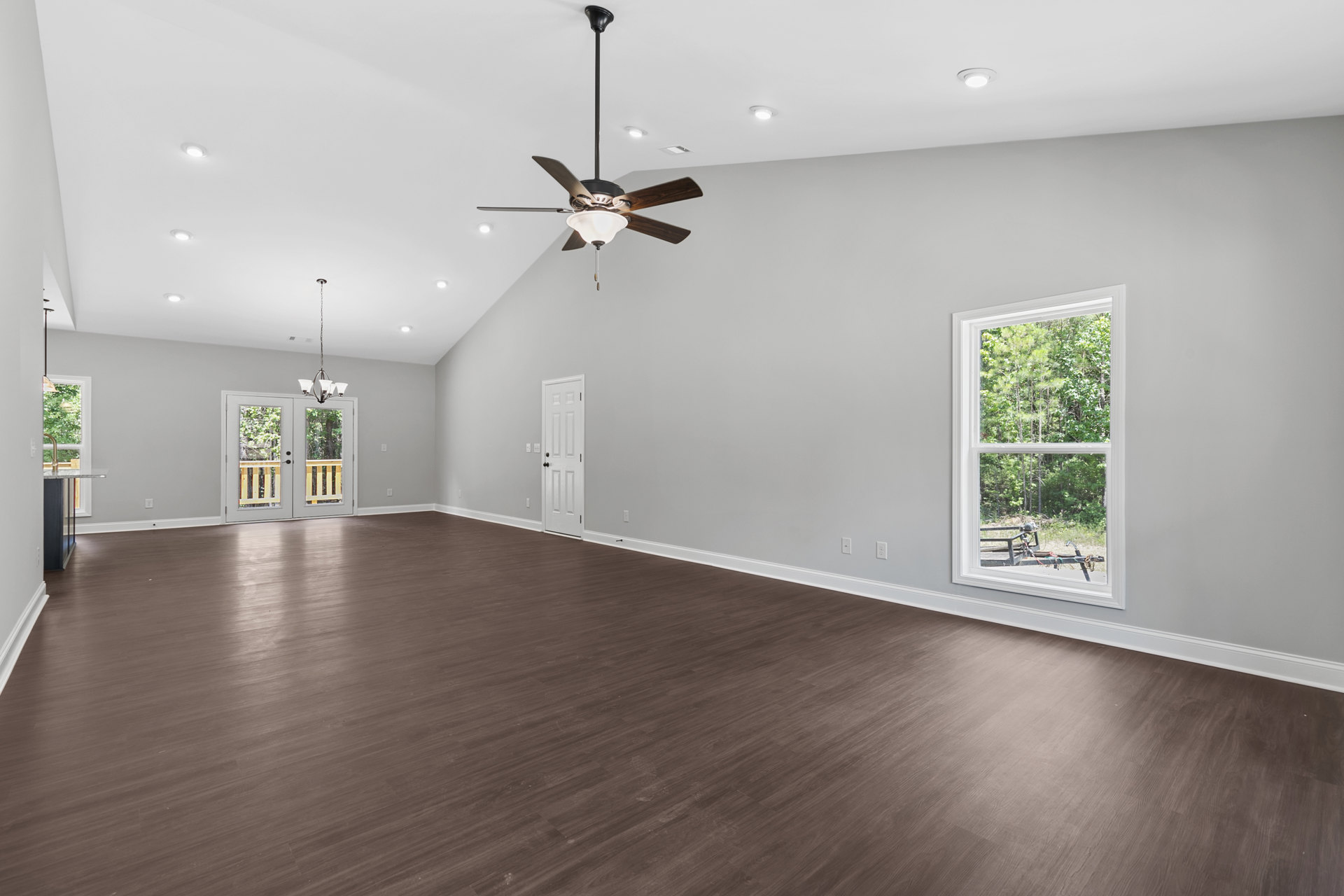 Spacious room featuring wood laminate flooring, white walls, ceiling fan with light fixture, double glass-paneled doors, and large window overlooking trees.