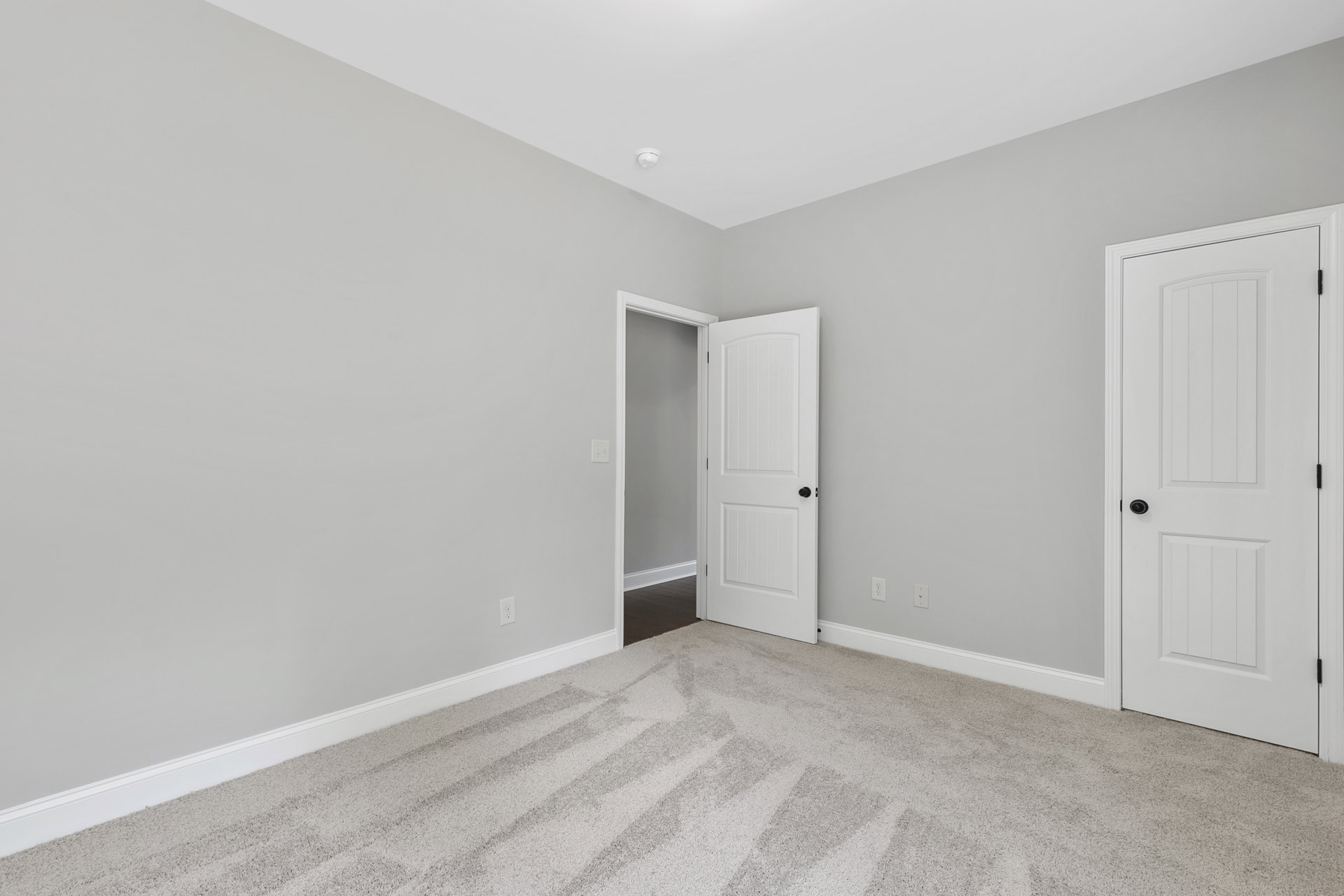 White carpeted room with open white door featuring black knob, white door frame and border, white ceiling with mounted camera.