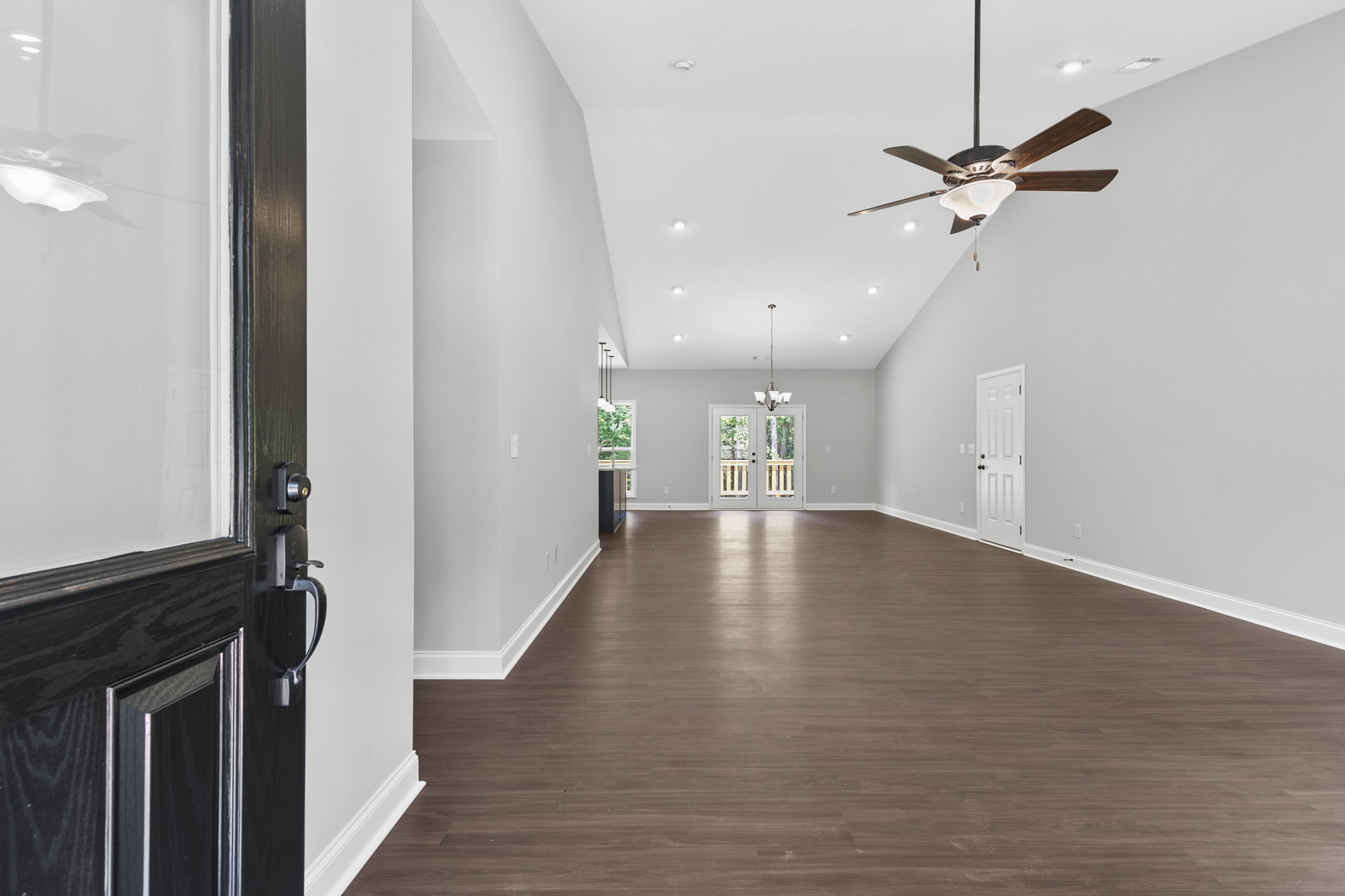 Spacious room with laminate wood flooring, white plaster walls, ceiling fan with light fixture, double white doors featuring black handles, and a chandelier above the entryway