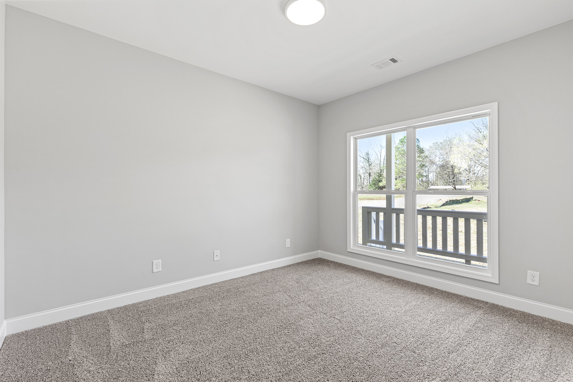 Carpeted room with large window and glass door opening to balcony, white walls, outdoor view of deck, trees, and fenced grassy area