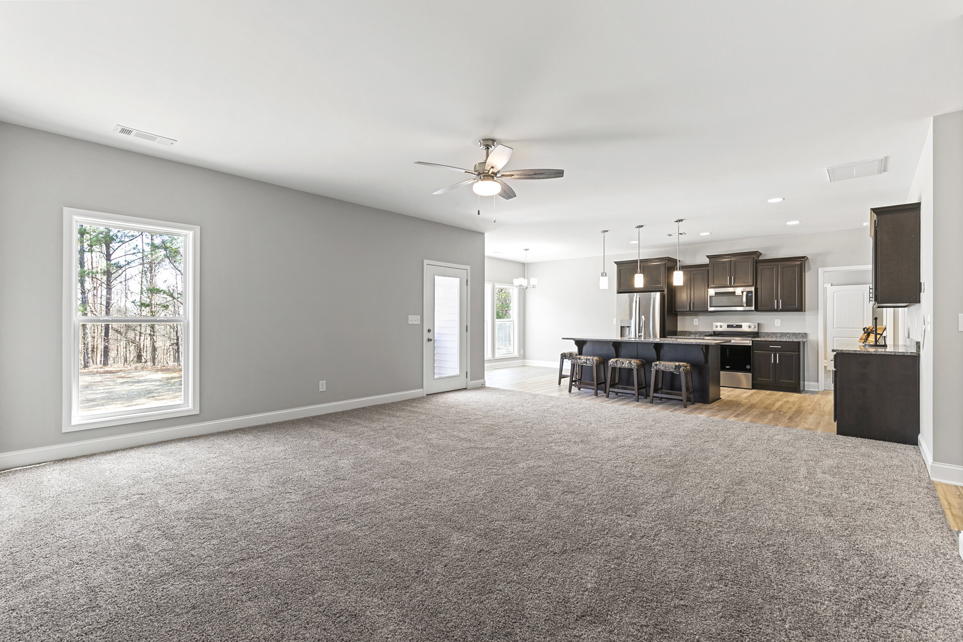 Open-concept kitchen and dining area with wood cabinetry, laminate flooring, white door with blinds, large window overlooking trees, camouflage-patterned stool, and neutral carpet