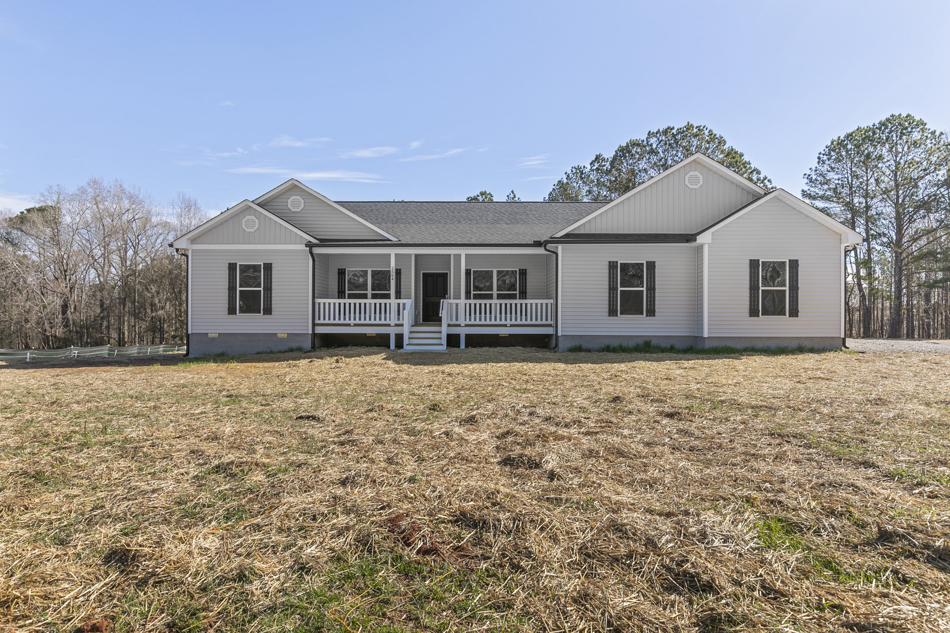 White two-story house with black front door, wide covered porch featuring white railings and chairs, surrounded by expansive green lawn and mature trees.