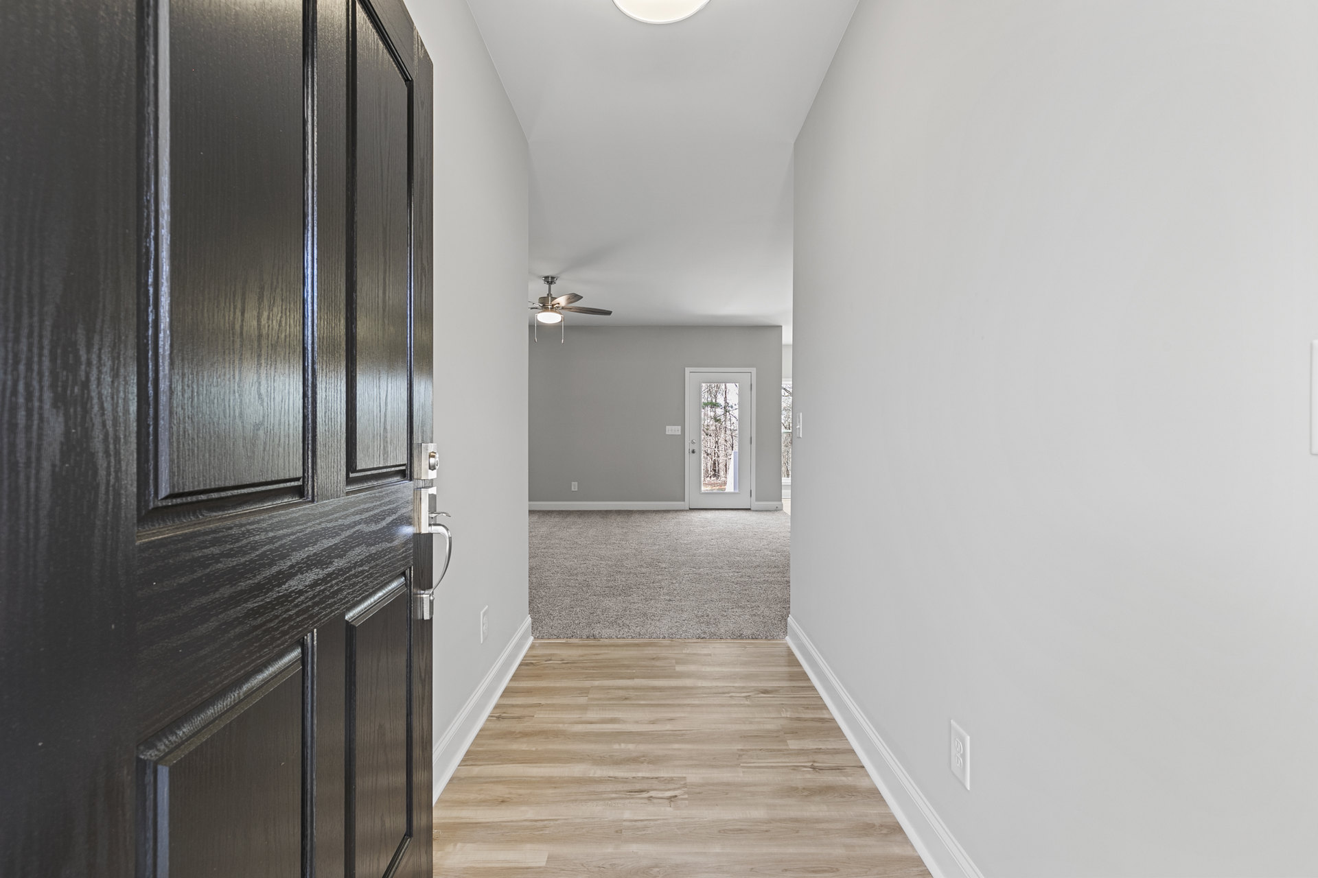 Hallway with white walls, wood flooring, white baseboards, black door, ceiling fan with light, and view of trees through the door