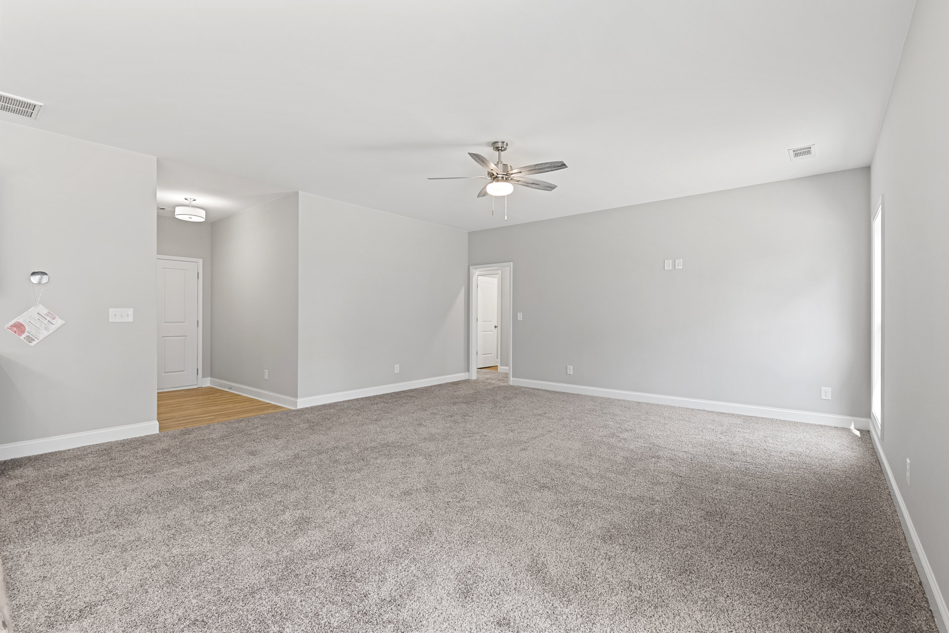 Spacious carpeted room with white walls, ceiling fan with light fixture, and open white door featuring a silver handle