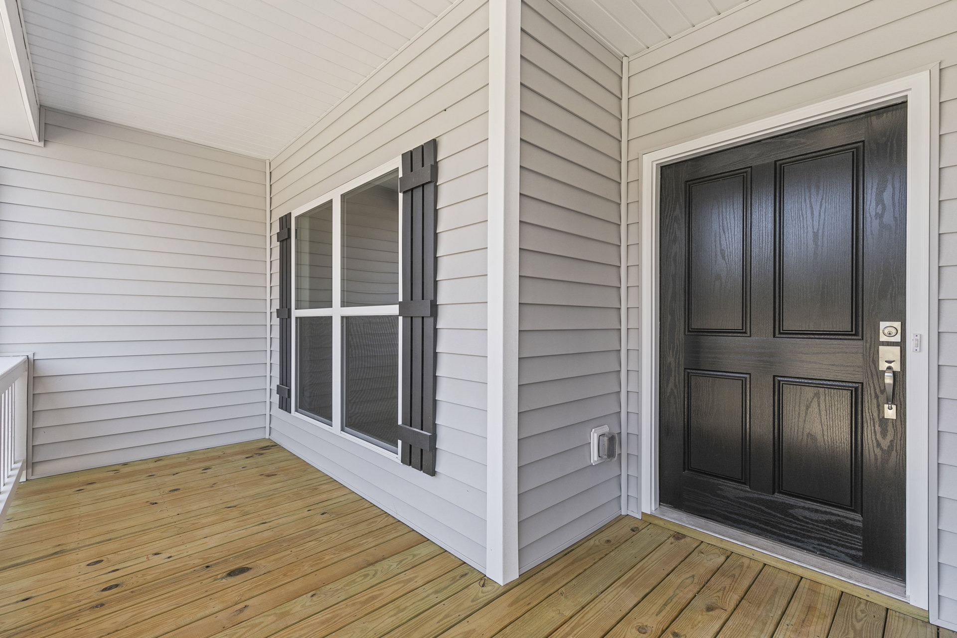Front porch with wood flooring, black door featuring silver handle, white siding, and large window adjacent to entry.