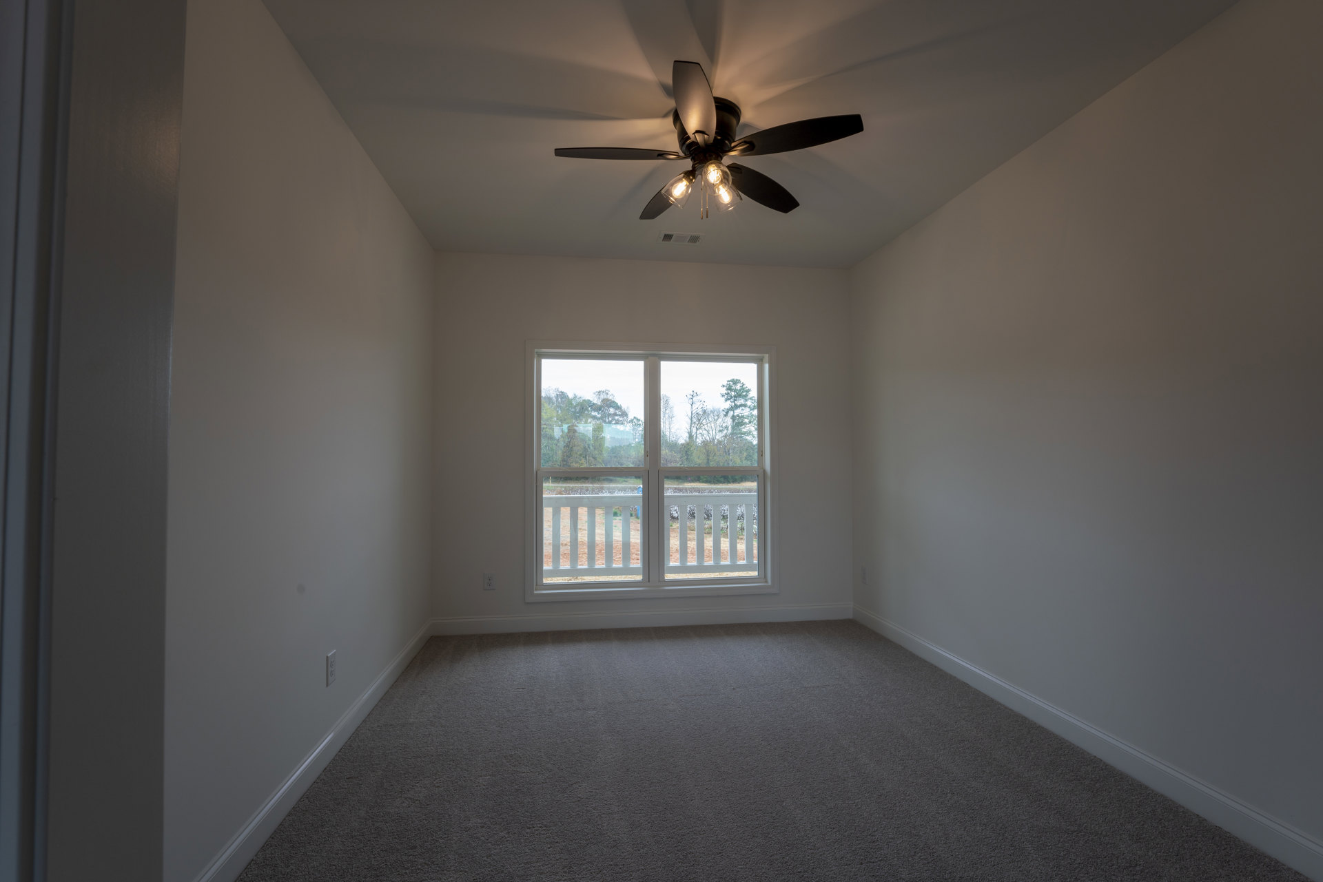 Ceiling fan with light fixture mounted on white plaster ceiling above carpeted floor; large window reveals view of trees, grass, and fenced field outside.
