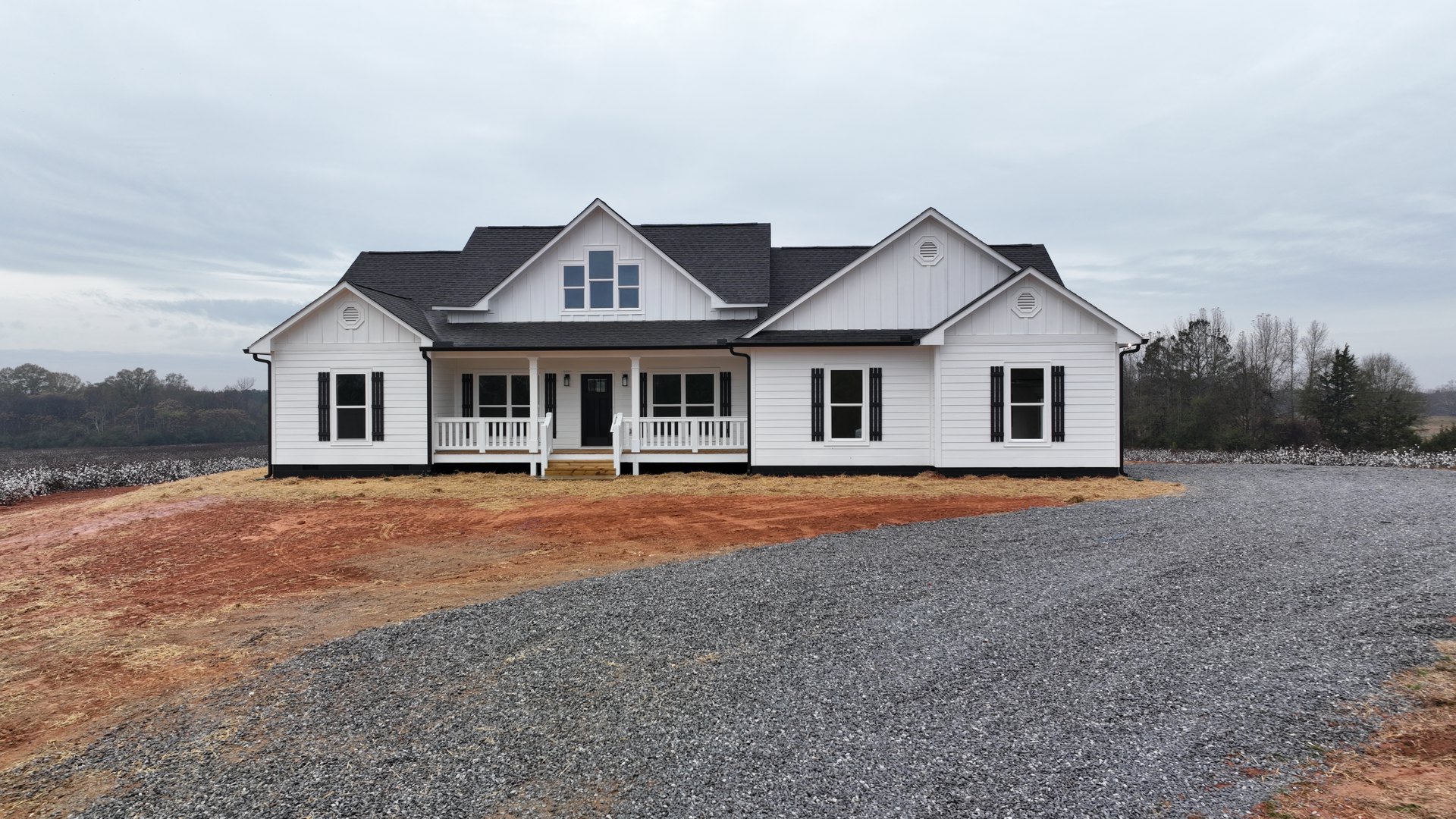 White house with black roof, gravel driveway, white deck railing, and white-framed windows surrounded by trees under a partly cloudy sky