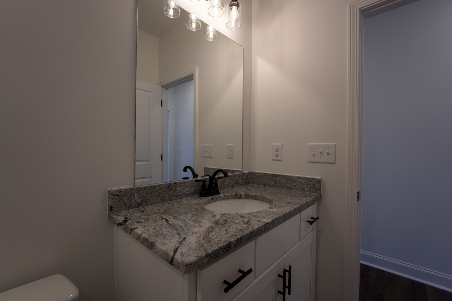 Bathroom with marble countertop, rectangular mirror above sink, white walls, black door, chrome faucet, row of switches on wall, tile flooring
