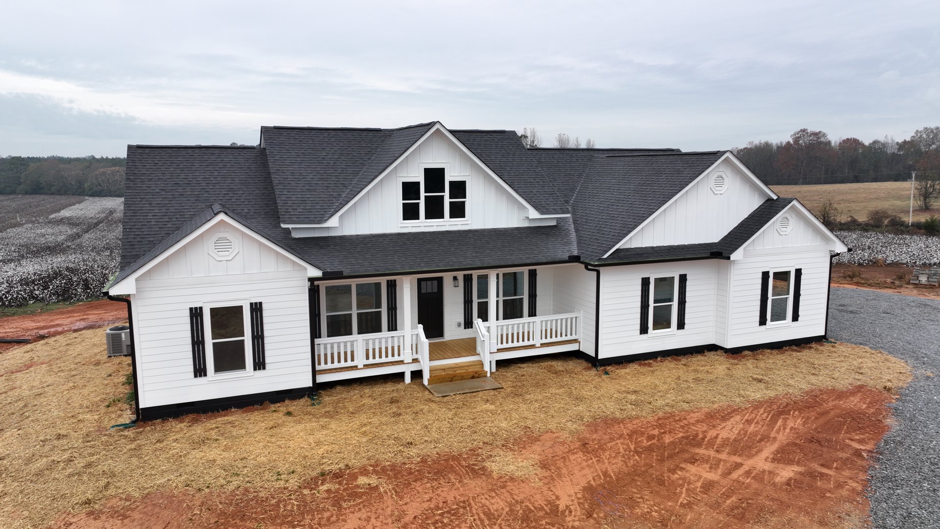 Two-story house with gray siding, black-trimmed windows, covered front porch, white railings, and stairs leading to a landscaped green yard under a cloudy sky