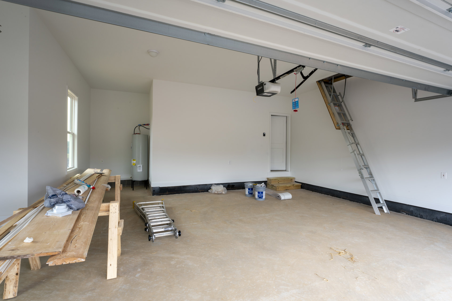 Unfinished room with exposed plaster walls, ceiling beams, two ladders, wood planks with tools, concrete floor, and building insulation visible