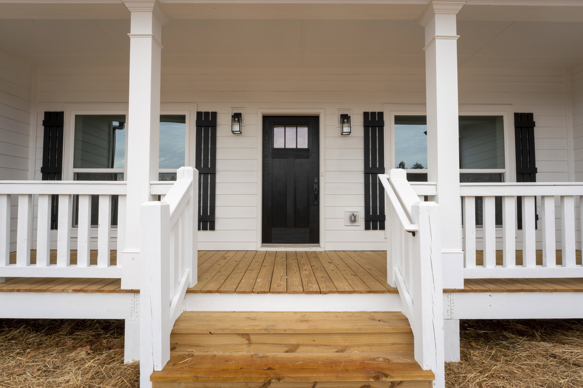White siding house with black front door, glass window insert, wooden deck and railing, outdoor stairs, and porch balusters