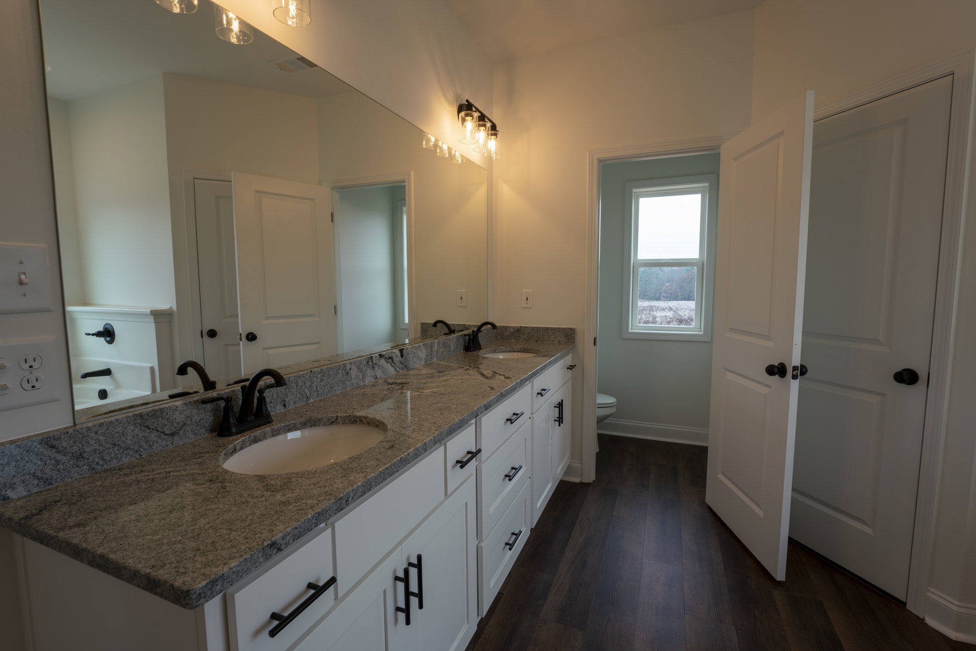 Bathroom with marble countertop, wood floor, black faucet, white door with black knobs, window overlooking trees and cotton field