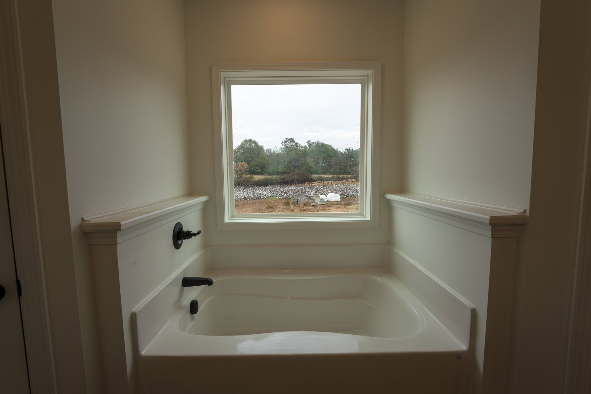 White freestanding bathtub beneath large window overlooking grassy field and cotton, black faucet mounted on wall, sunlight streaming in, trees and fence visible outside