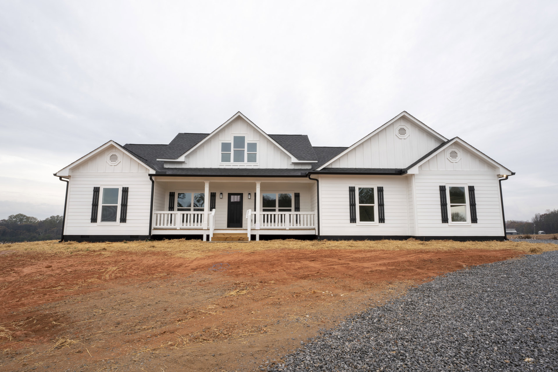 White siding house with black shutters, black front door with window, white porch railing, gravel and dirt landscaping, cloudy sky overhead