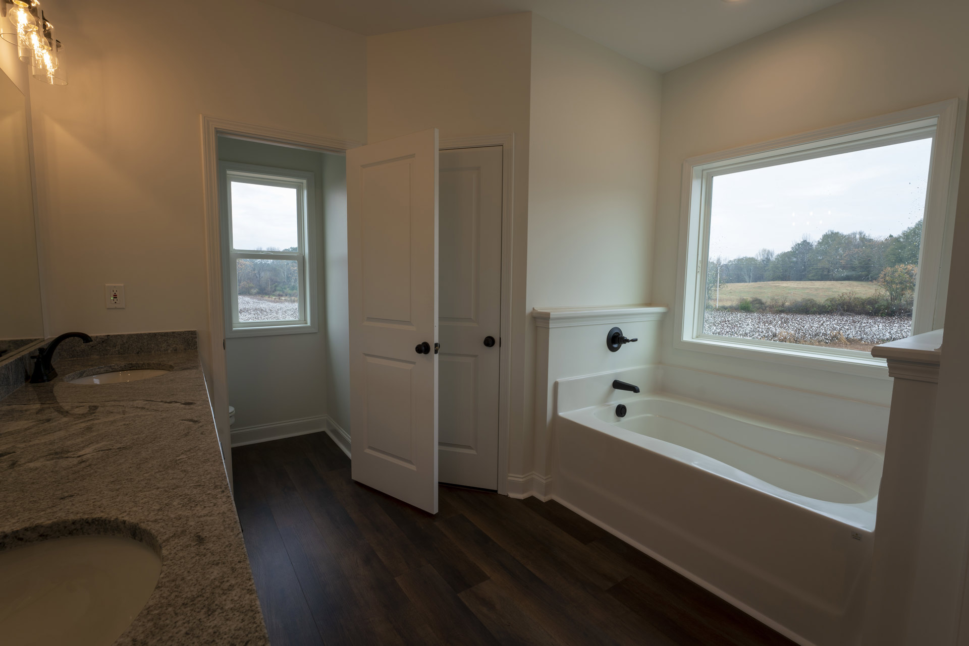 Freestanding bathtub beneath large window with tree views, white bathroom walls, stone countertop with undermount sink, light wood flooring