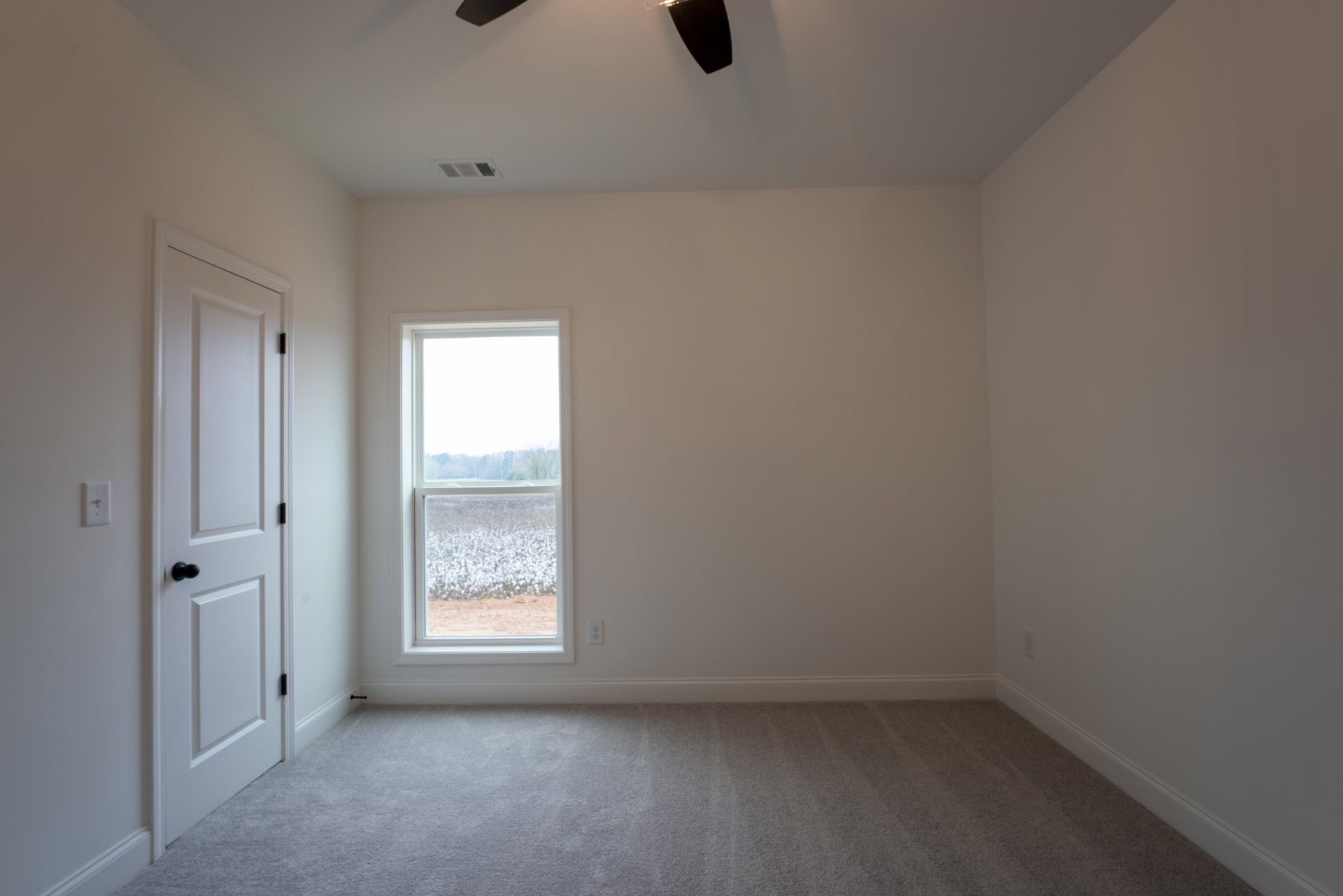 Ceiling fan mounted on white ceiling above carpeted floor, white baseboards, white door with black knob, multiple windows showing snowy field, cotton field, trees, and blue sky.