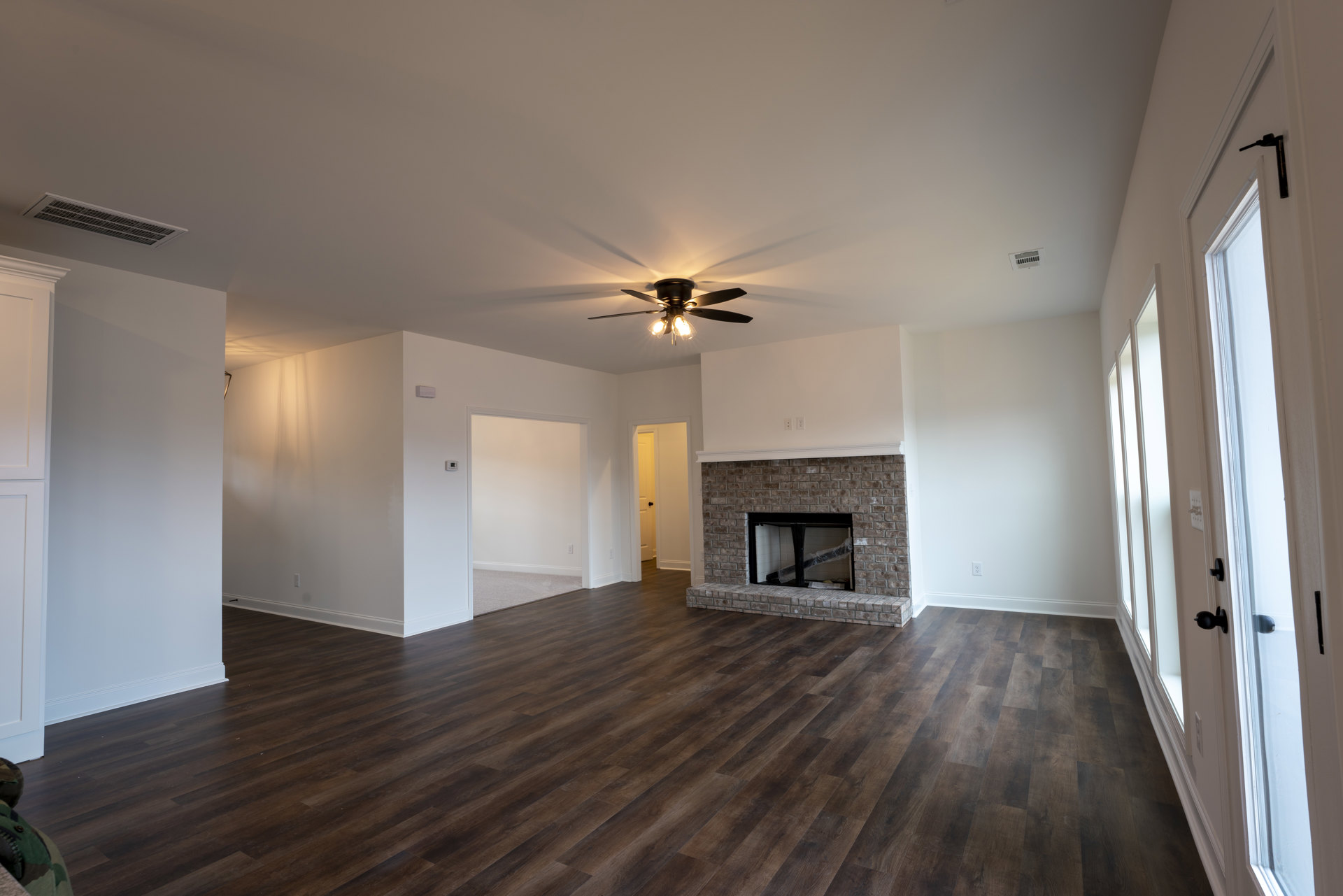 Living room with brick fireplace featuring black metal frame, hardwood flooring, ceiling fan with lights, black metal shelf, and stuffed animal on floor