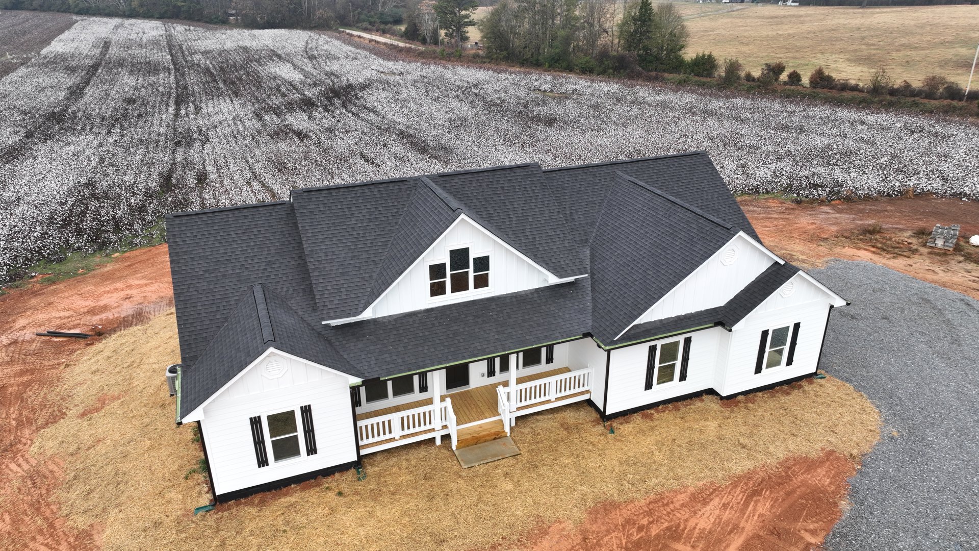 Large white house with black roof, multiple windows, white porch with wood railing, pile of bricks on pallets in front, expansive cotton field surrounding property.
