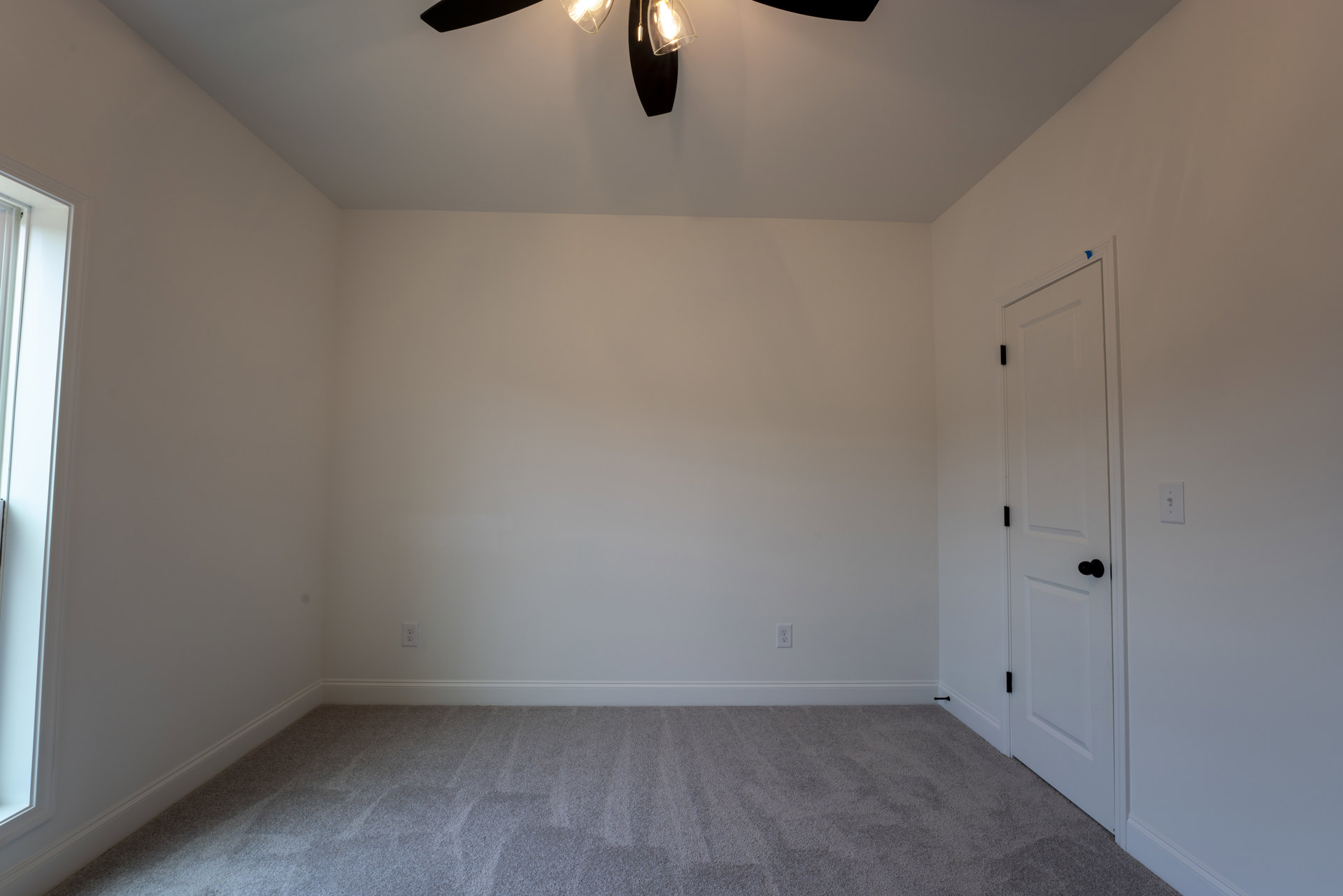 Ceiling fan with dark blades mounted on white plaster ceiling, carpeted floor, white door with black knob, clear glass light fixture, and large window with trim in a bright