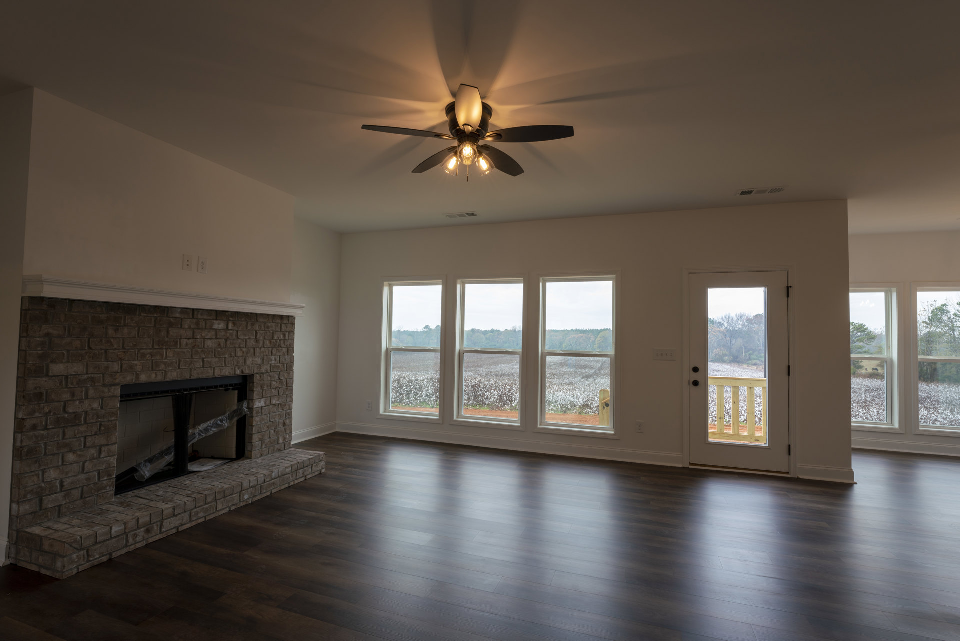 Living room with hardwood floors, brick fireplace wrapped in plastic, black metal frame, ceiling fan with lights, large window overlooking cotton field