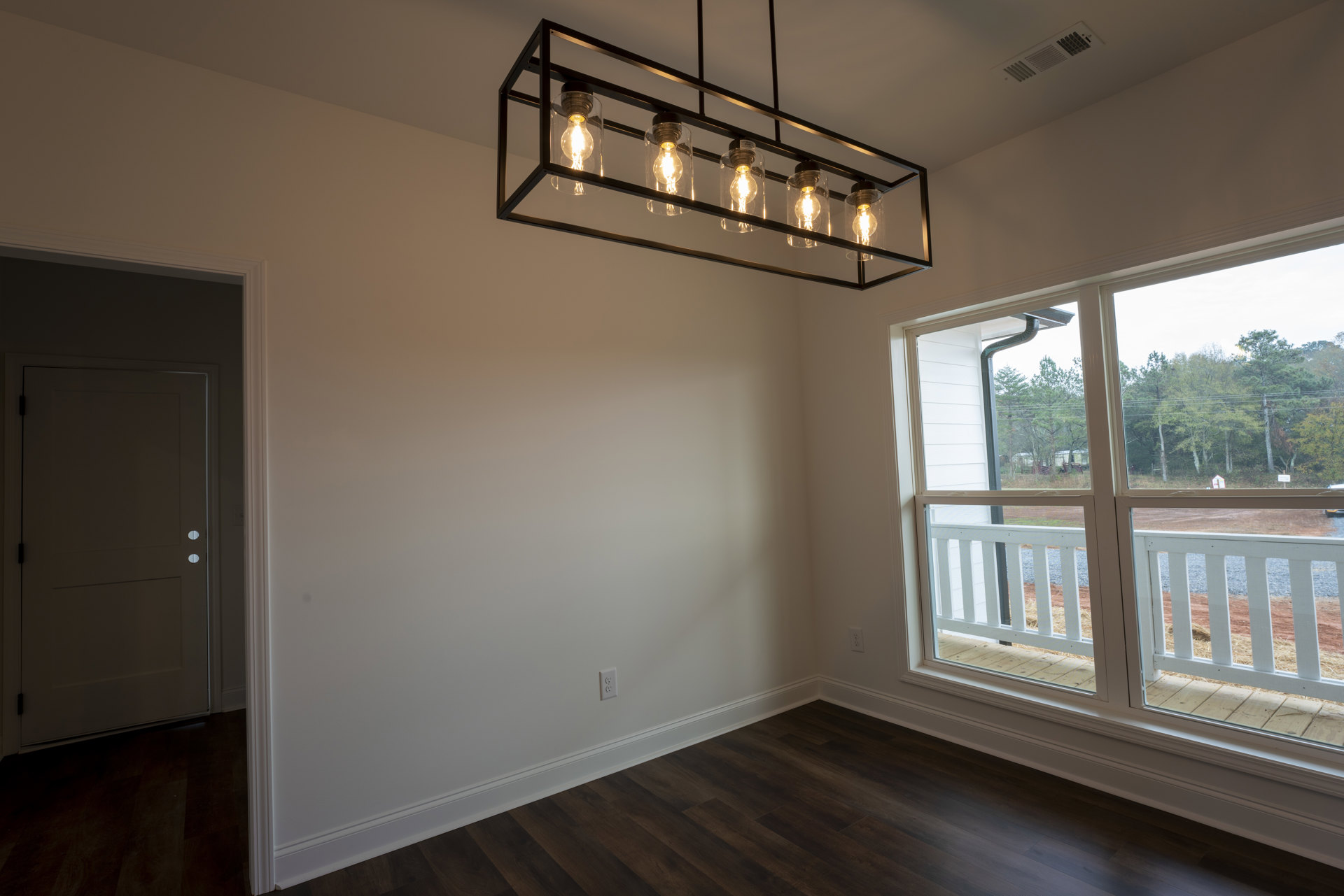 Black bar ceiling light fixture with exposed bulbs, white railing and fence, white wall, door featuring round holes, wood flooring, large window providing daylight.