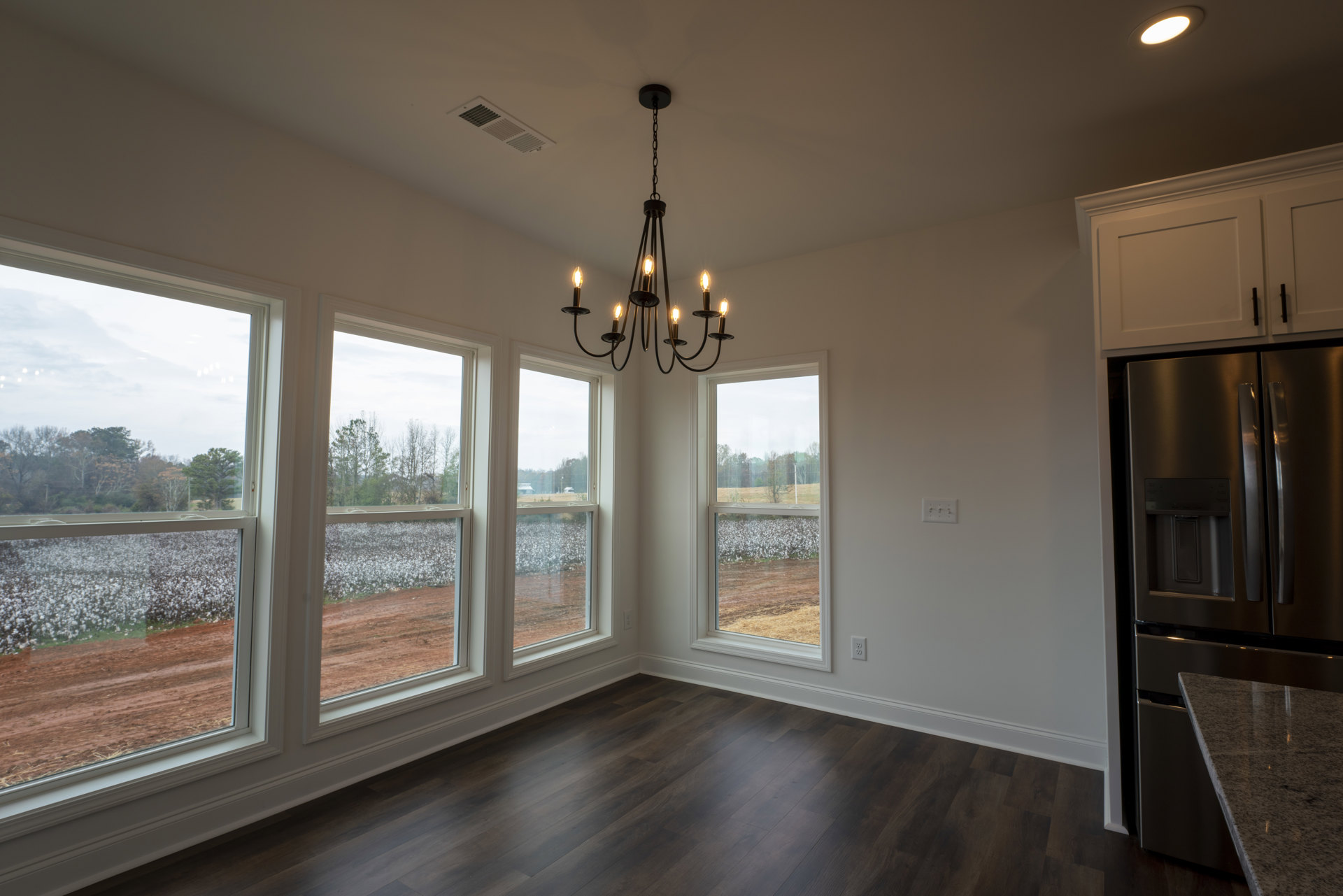 Chandelier with glass shades hanging from a white ceiling, large windows overlooking a cotton field, laminate wood flooring, neutral painted walls, stainless steel refrigerator