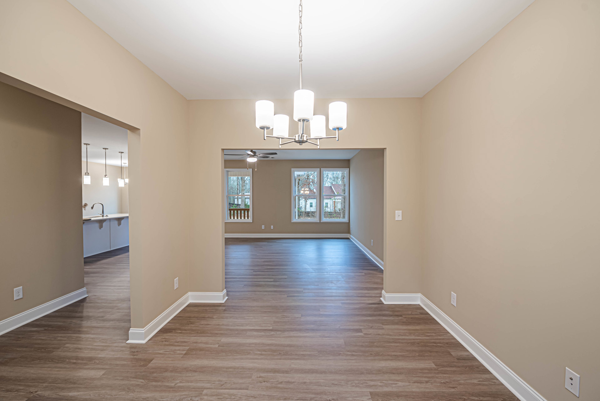 Open room with wide-plank wood flooring, white plaster walls, large window overlooking trees, ceiling-mounted chandelier, and visible wall outlet.
