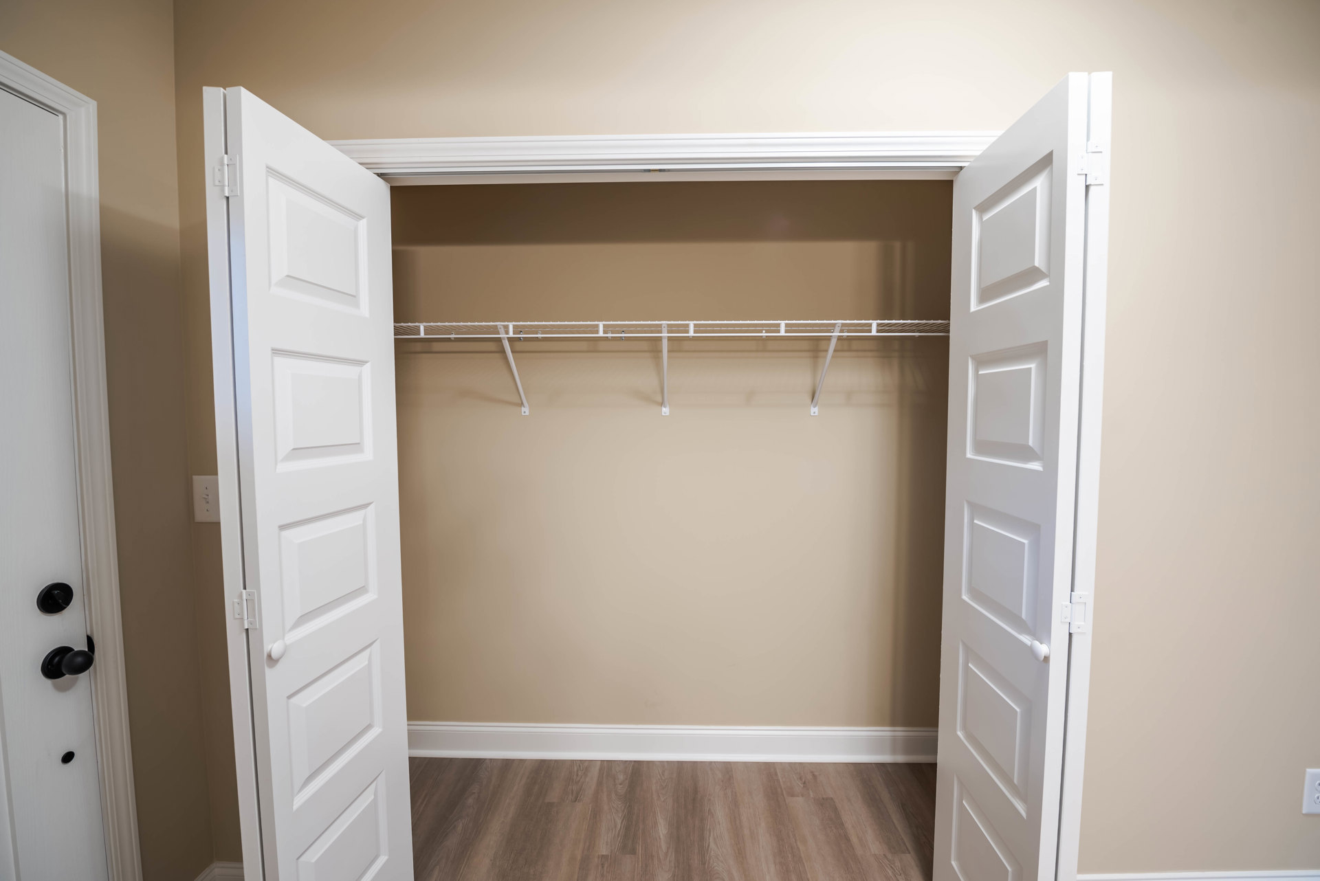 Closet with open white doors, wood floor with white baseboard, square panel doors with black knobs, white walls, white door frame, and light switch.