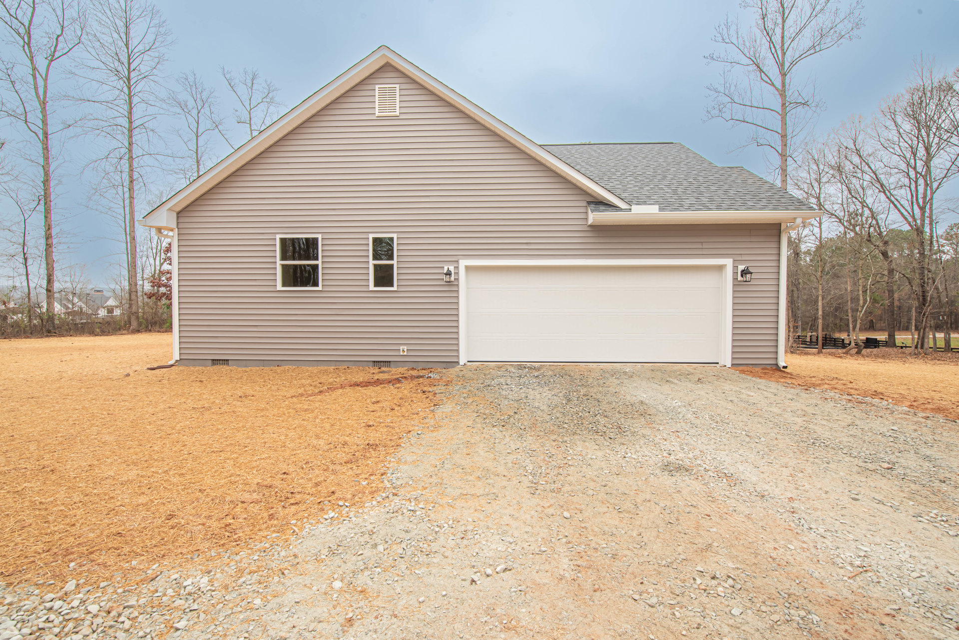 Grey siding house with brown roof, white garage door, white vent, and white-framed windows; concrete driveway bordered by lawn and trees.